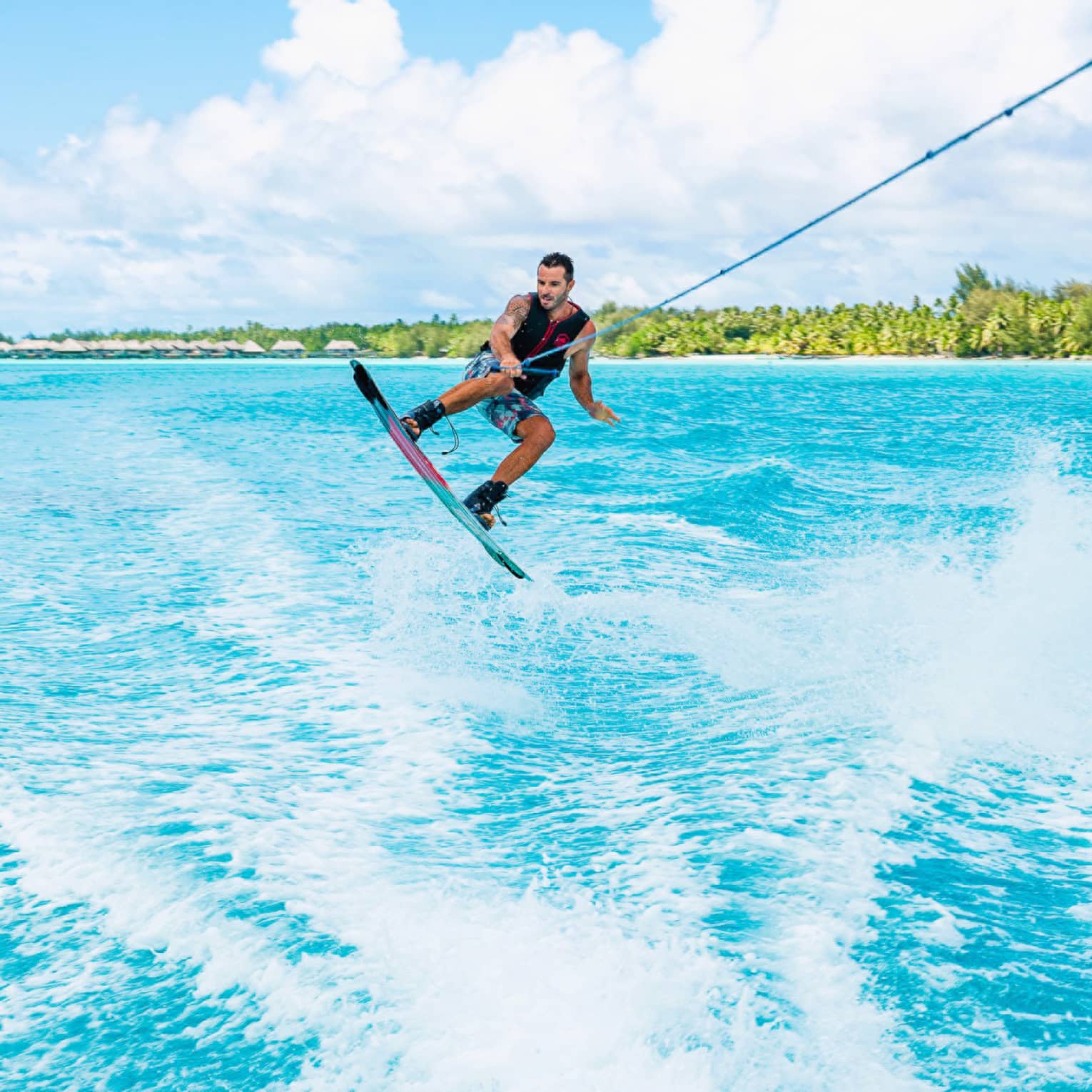 A wakeboarder leaps above the waves, one hand holding a taut rope as they're towed through the ocean beneath a sunny sky.