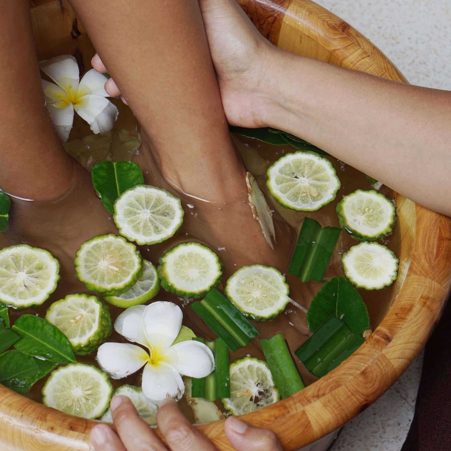 A guest receiving a foot massage at the resort spa in a wooden basin filled with water, lime slices, green leaves and white flowers.