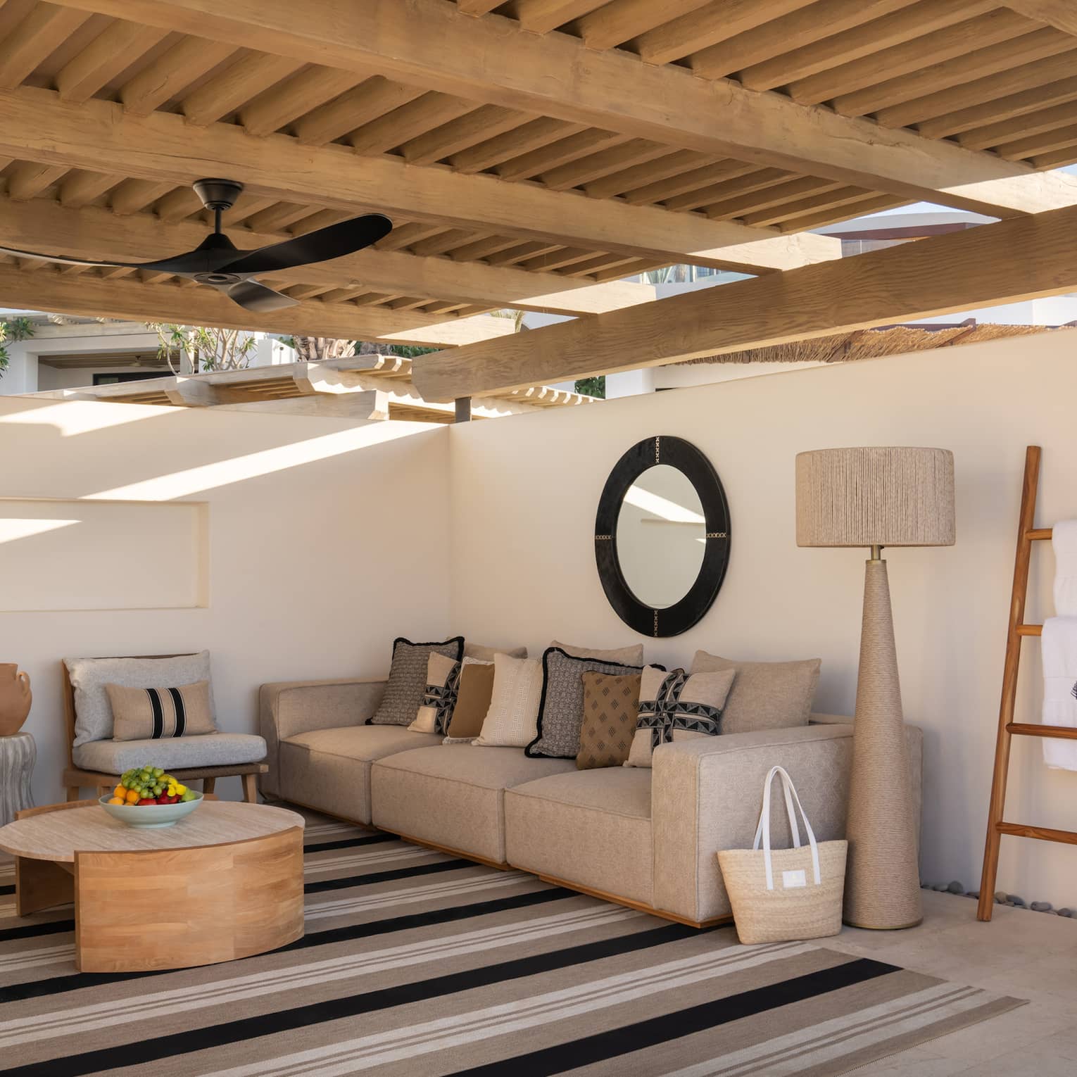 A serene cabana with a raised wooden roof and plaster walls. It's filled with comfortable furniture and a large striped rug.
