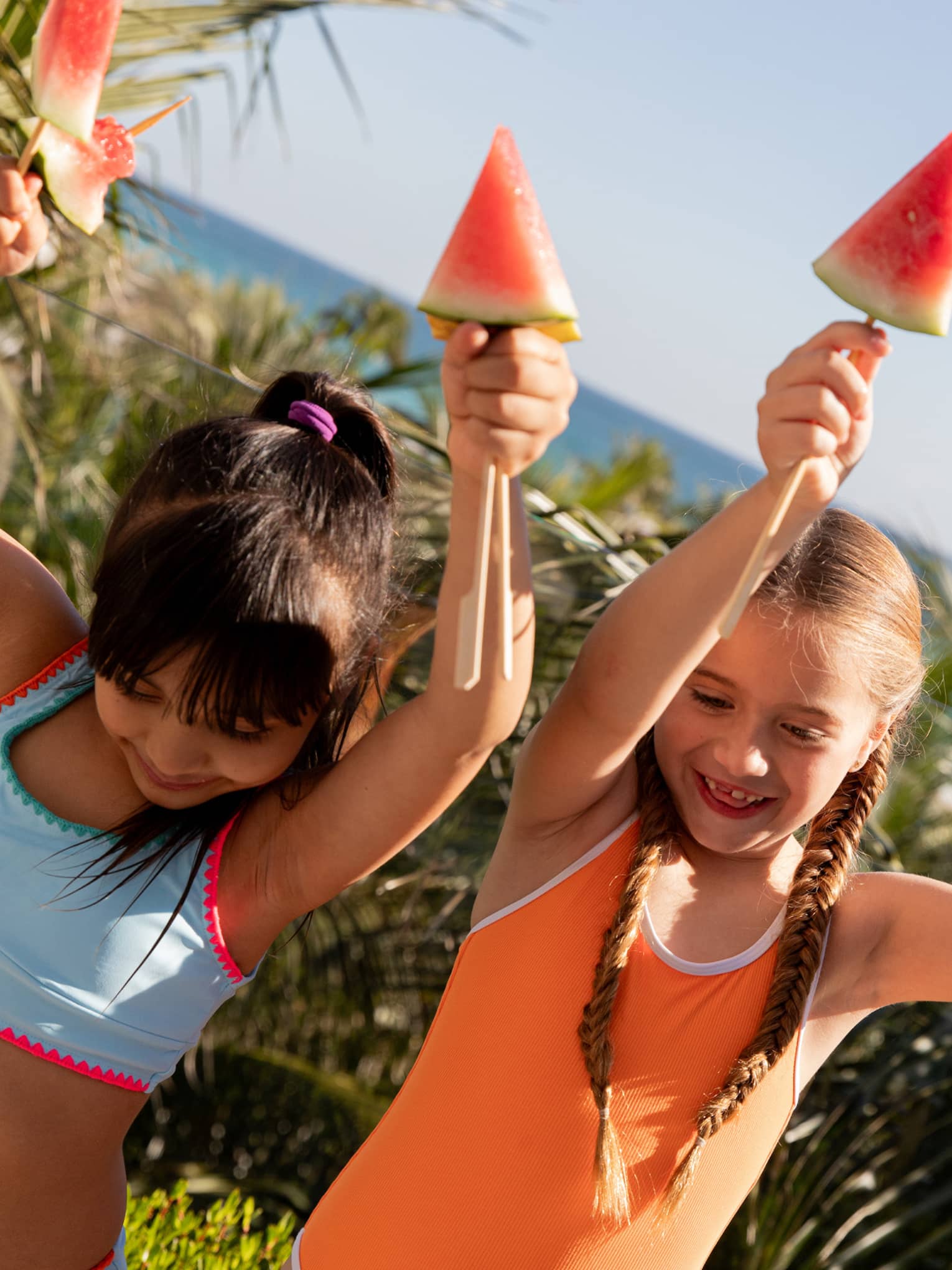 Two young children wearing bright orange and blue bathing suits smile while holding watermelon wedges above their heads