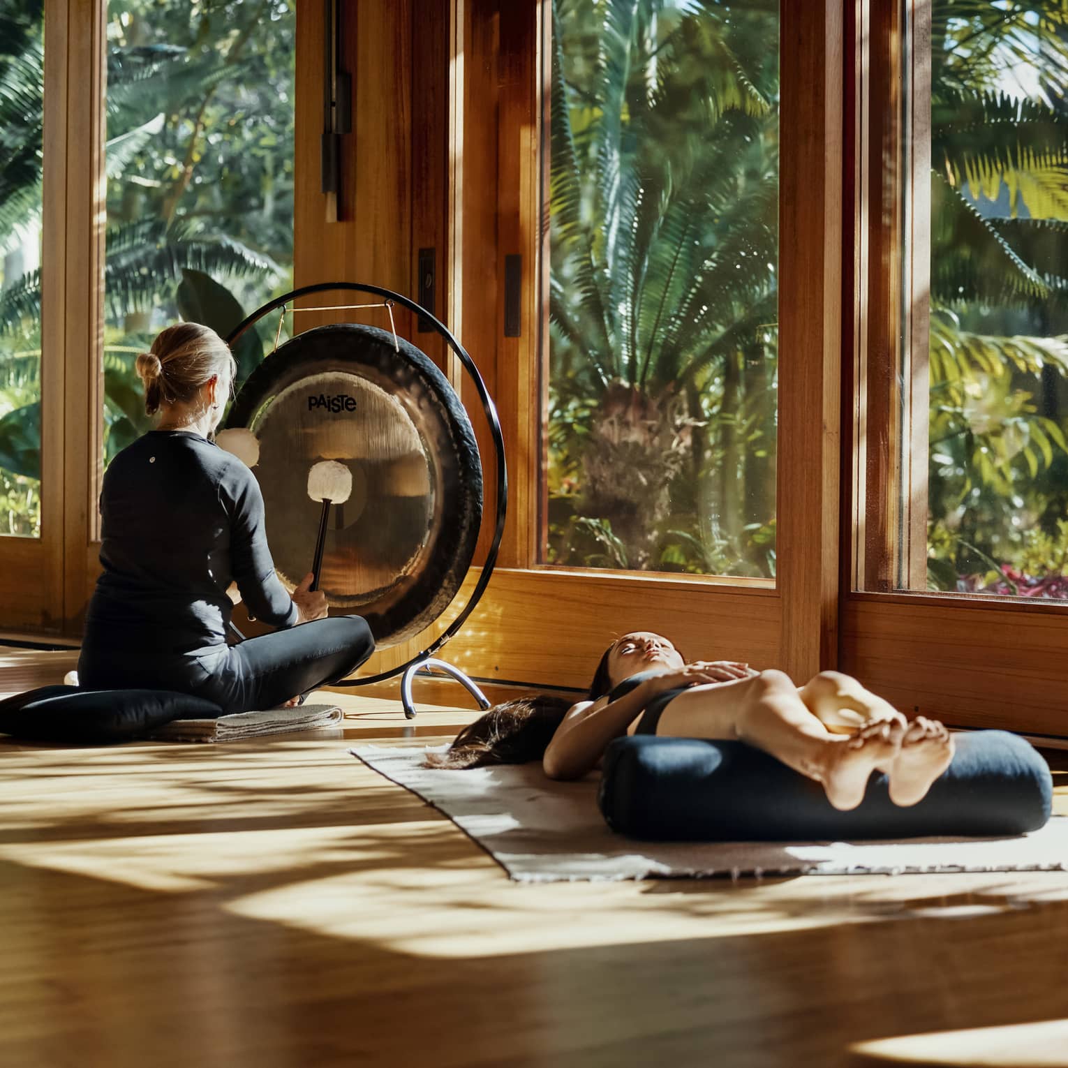 During a sound bath, a meditator lays on a mat, legs resting on a bolster, as a practitioner plays a gong with two mallets.