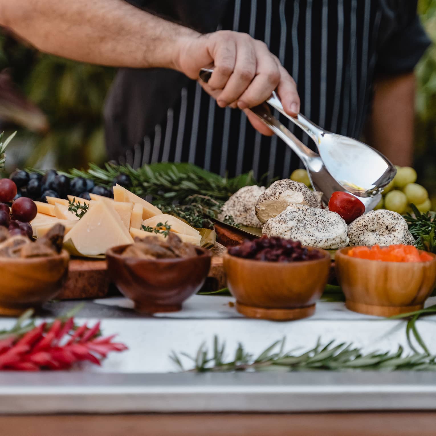 A chef's hand holds silver tongs over a display of grapes, dried fruits and cheese slices