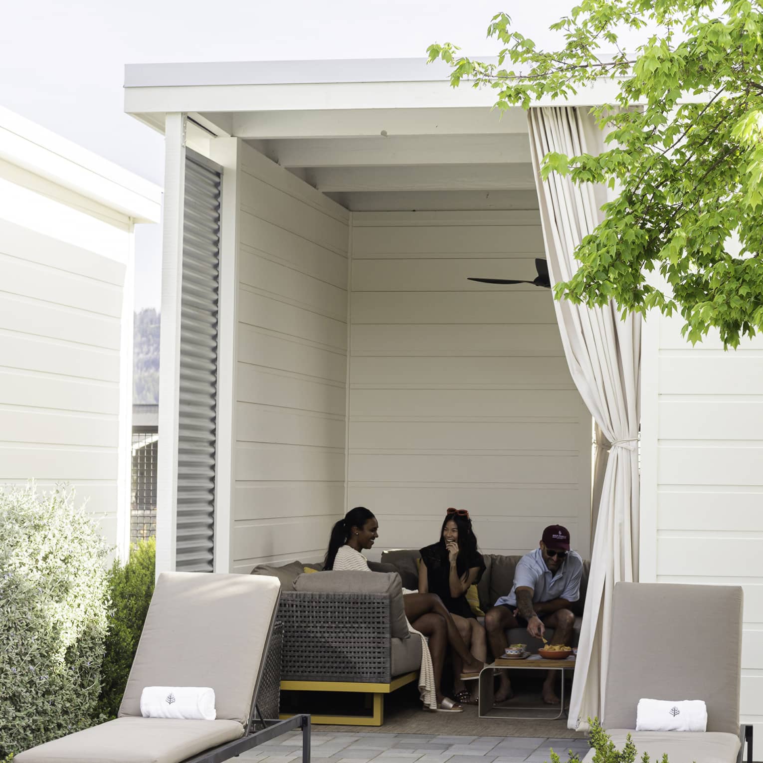 Three guests eat snacks on a large couch in a square cabana with a tied-off curtain and two lounge chairs at the entrance.