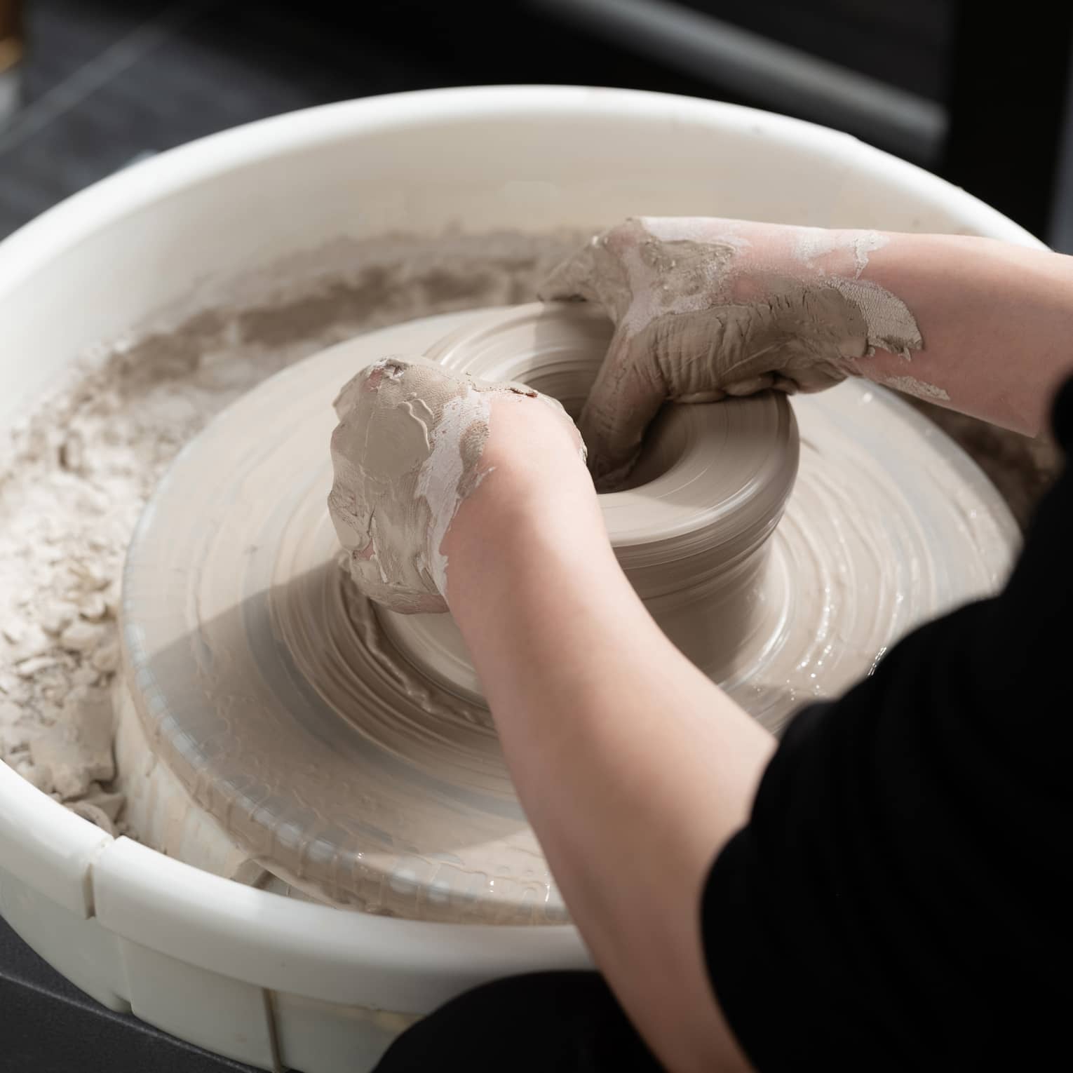 Close-up of two hands covered in wet beige clay shaping a pot on a spinning pottery wheel. The potter is wearing black.