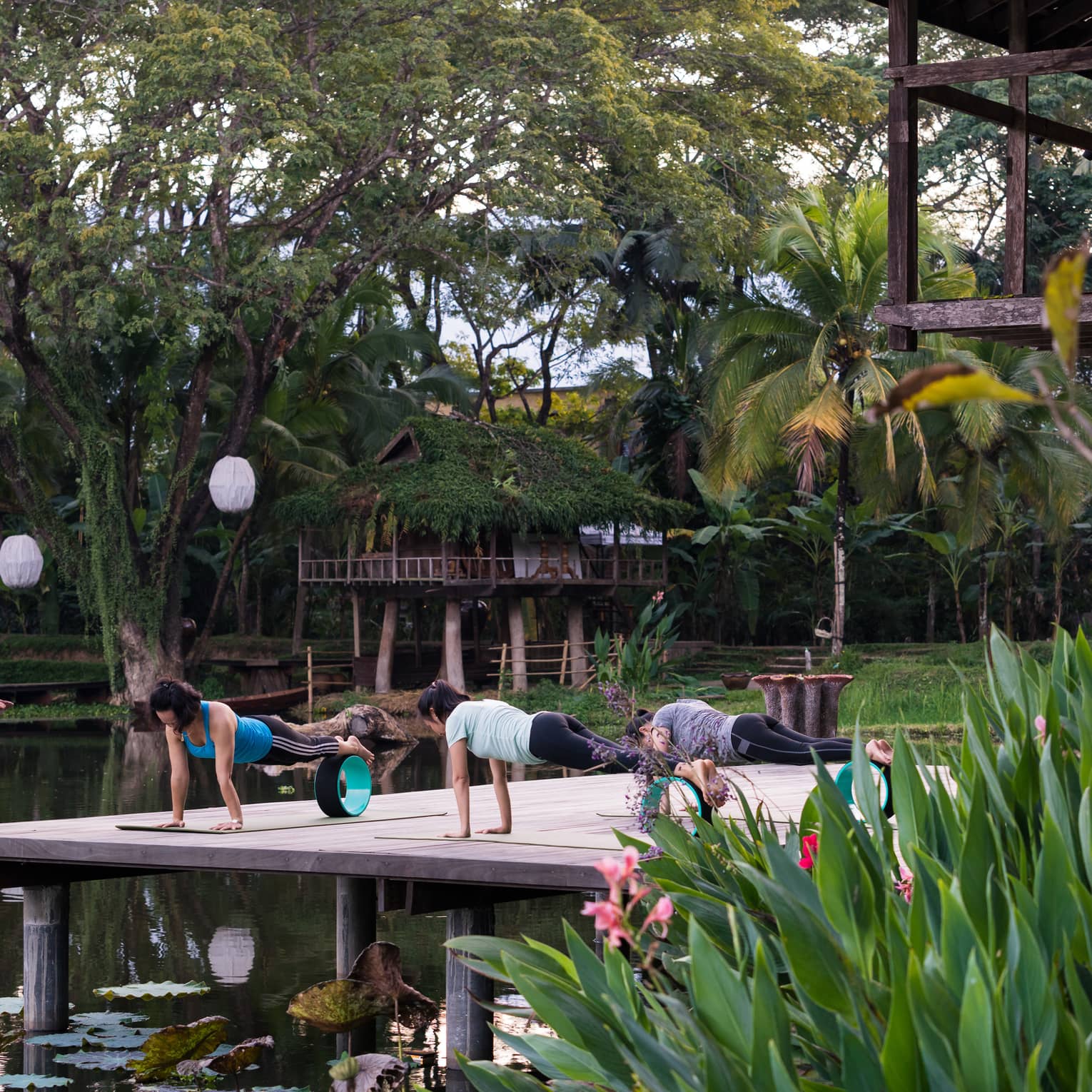 On a wooden deck above the water, a woman leads two people in a plank yoga move, with the feet balancing on a wheel