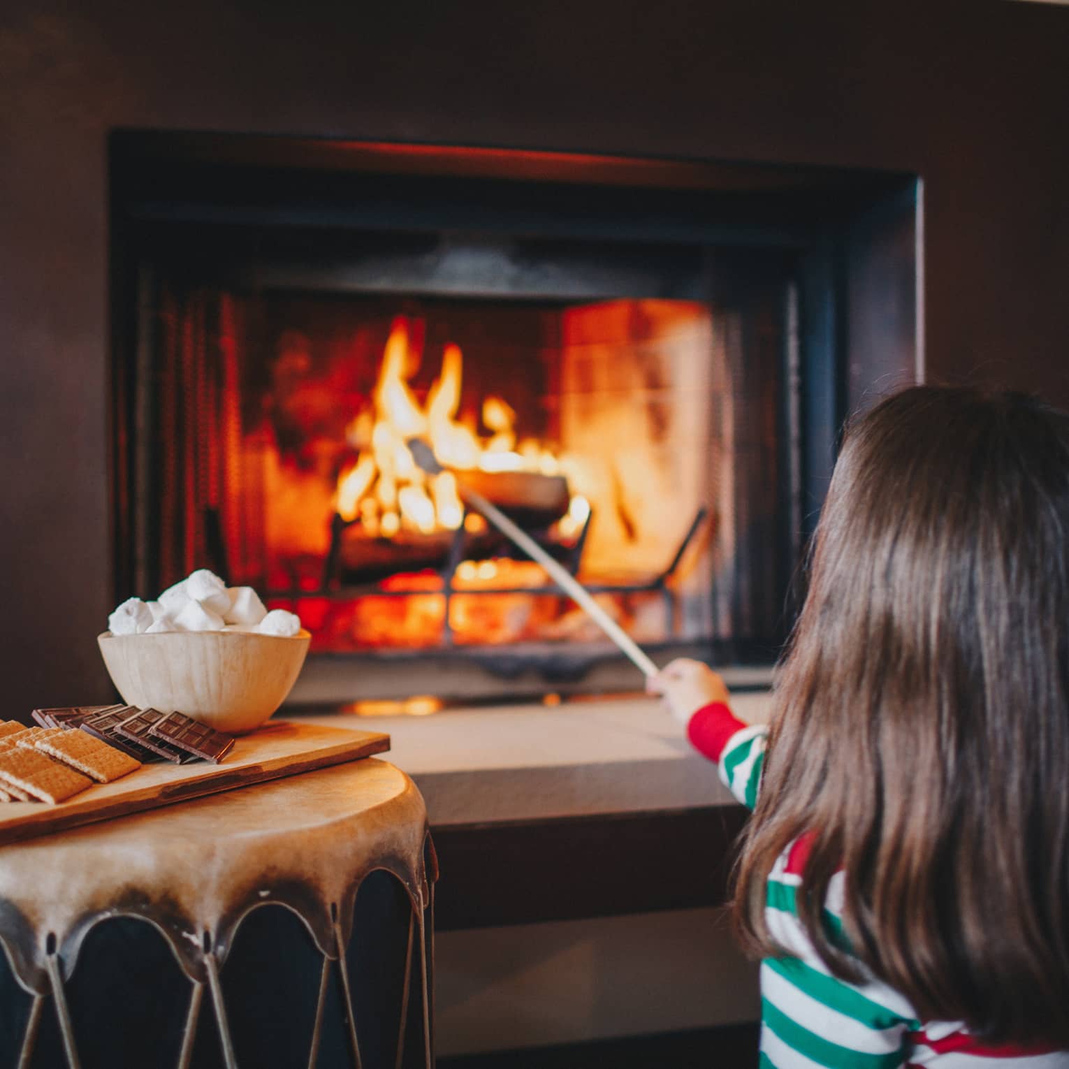 A little girl in red and green pajamas roasts marshmallows in a fireplace