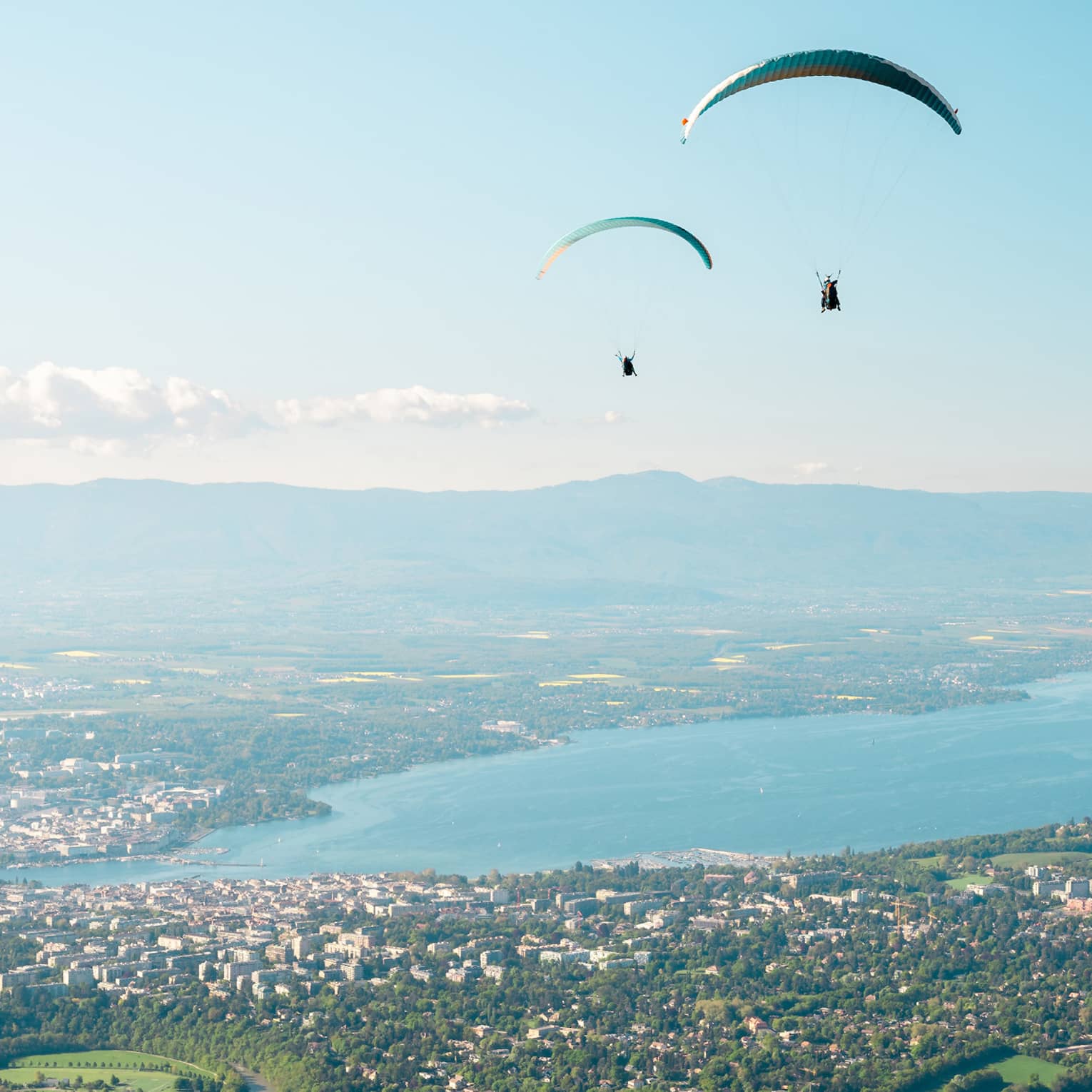 Paragliders soaring above a vast landscape with a lake, green fields and a city below, with distant mountains and a clear blue sky in the background