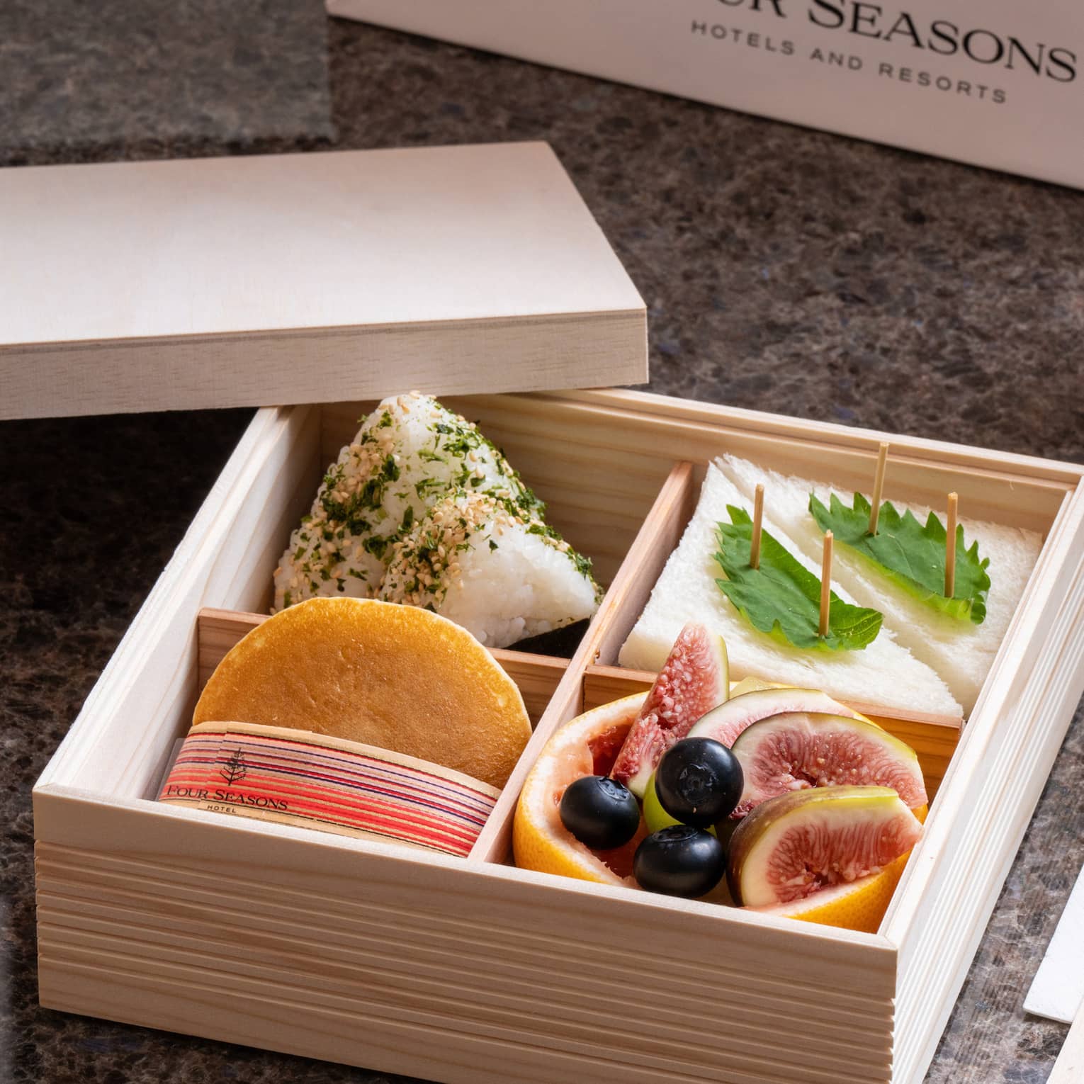 A wooden Four Seasons bento box with four compartments holding sandwiches, fruits and delicacies, beverage bottles nearby.