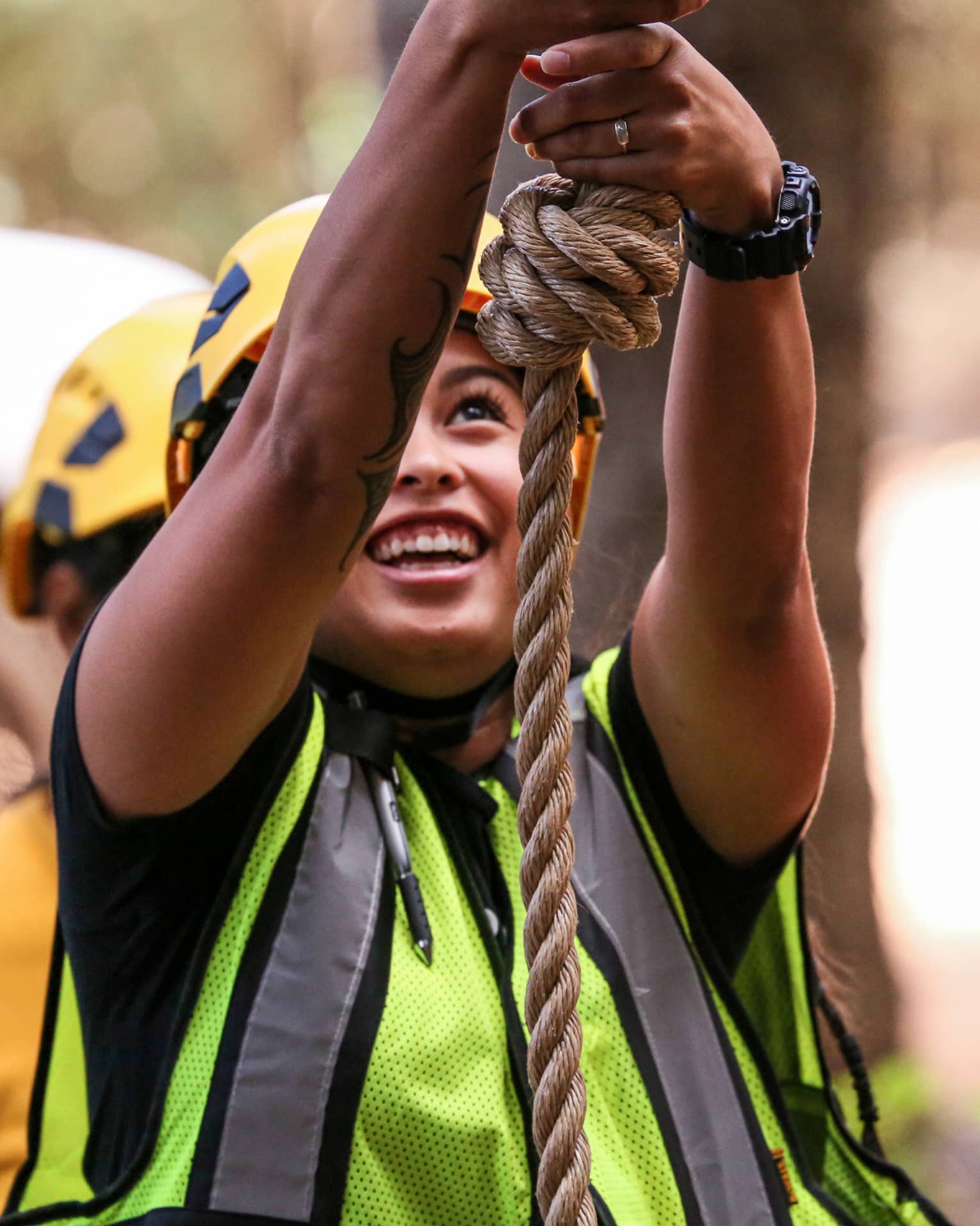 A woman in a helmet and safety vest smiles as she holds on to a rope