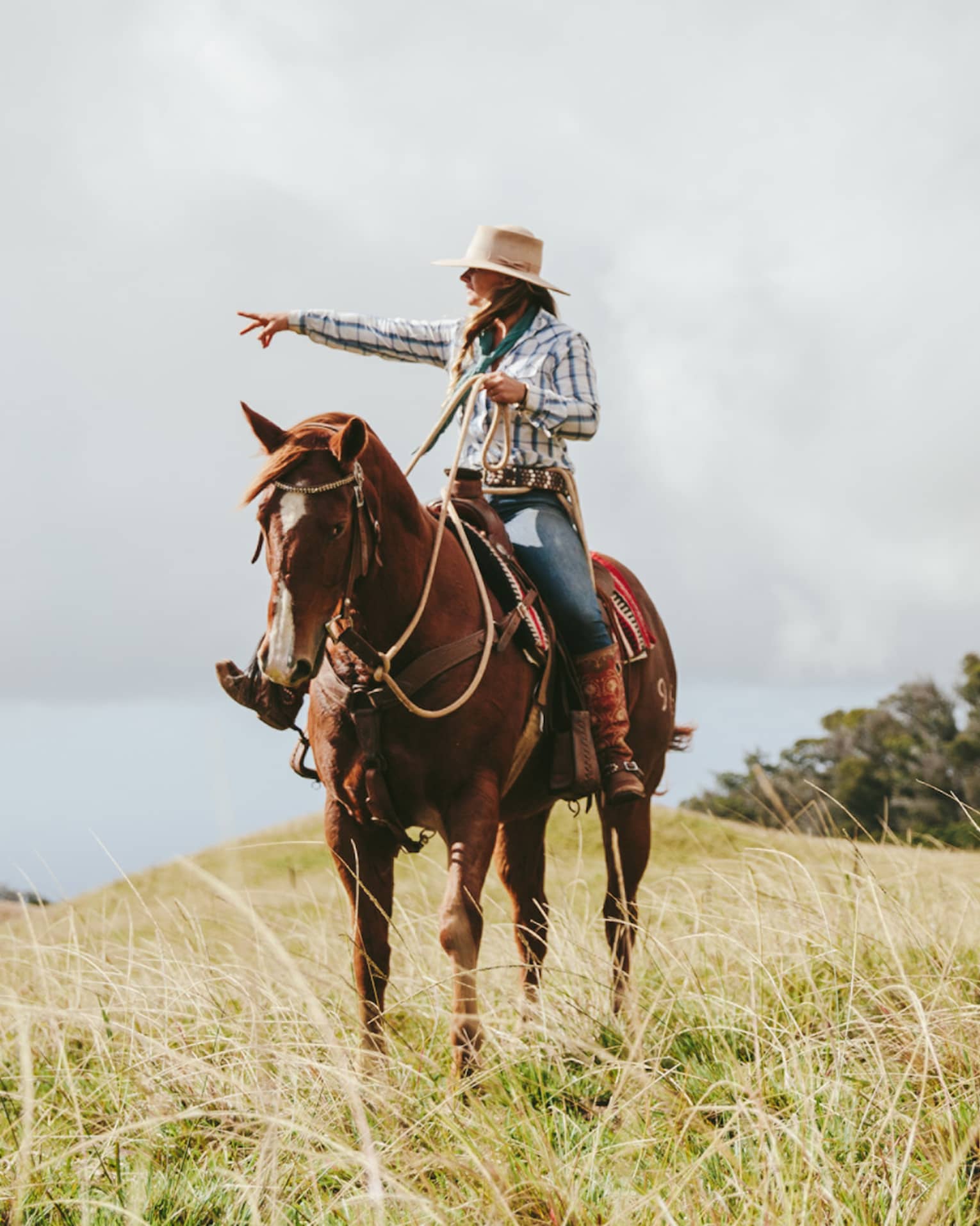 A couple riding horses, looking out at the long grass fields, with grassy hills and trees in the distance on a cloudy day.
