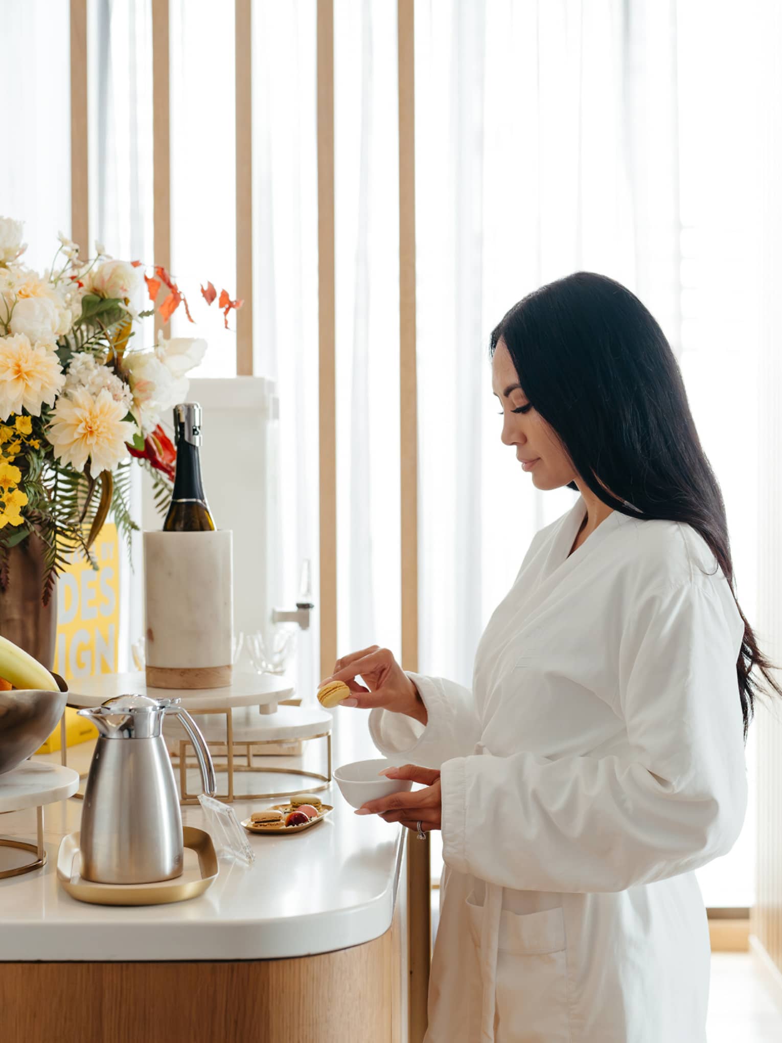 A guest in a white robe stands in a light-filled interior space in front of a counter holding a floral arrangement, a bowl of fresh fruit, a plate of small food items, a carafe and other beverage items.