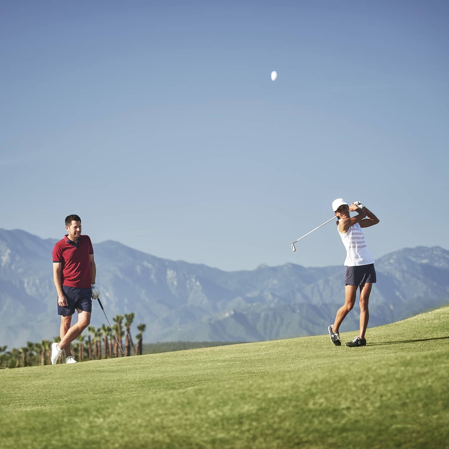 A golfer leans on their club observing another's swing on a picturesque golf course silhouetted by a mountain range.