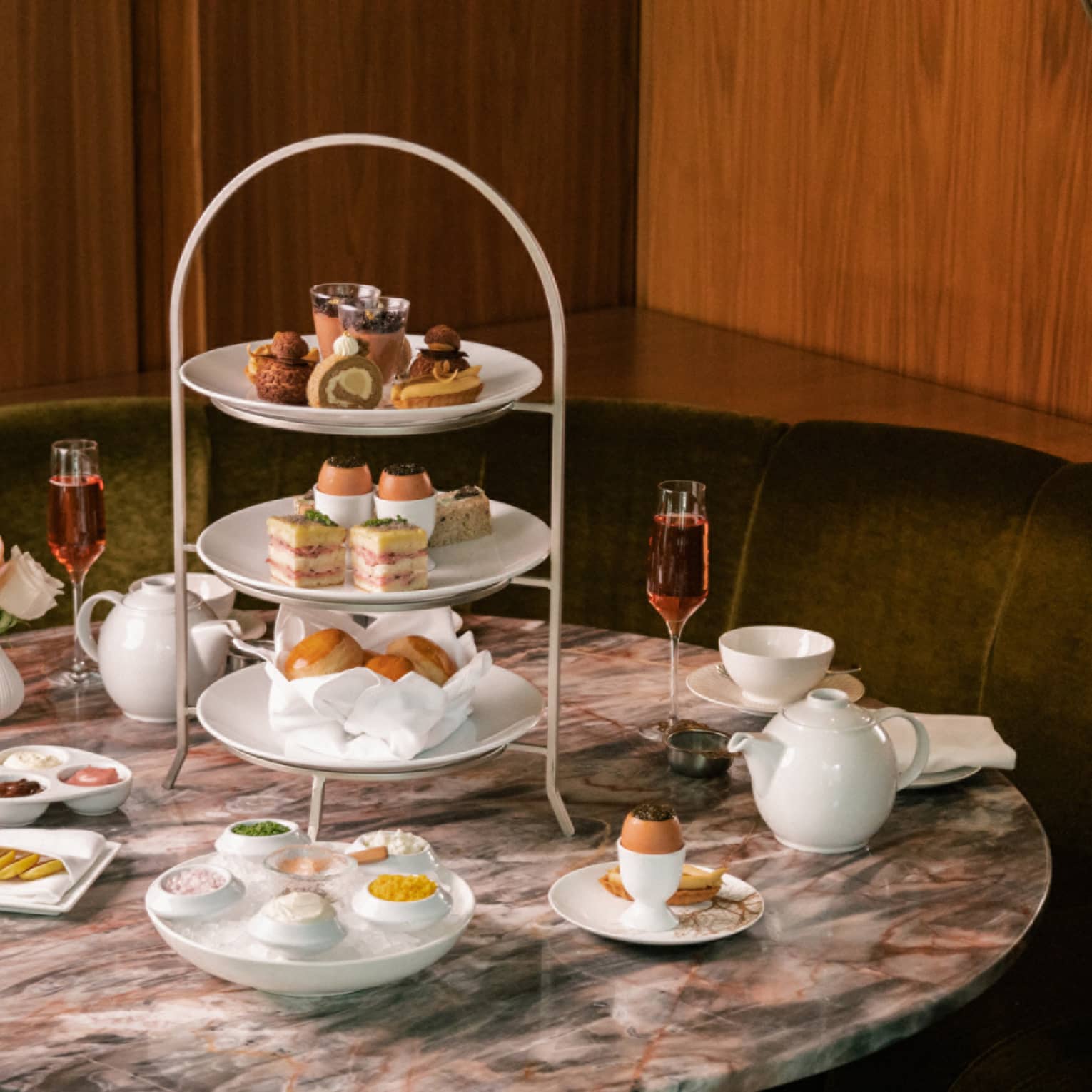 A round marble table set for tea with dainty pastries, two cocktail flutes and a three-tiered tray laden with delicacies.