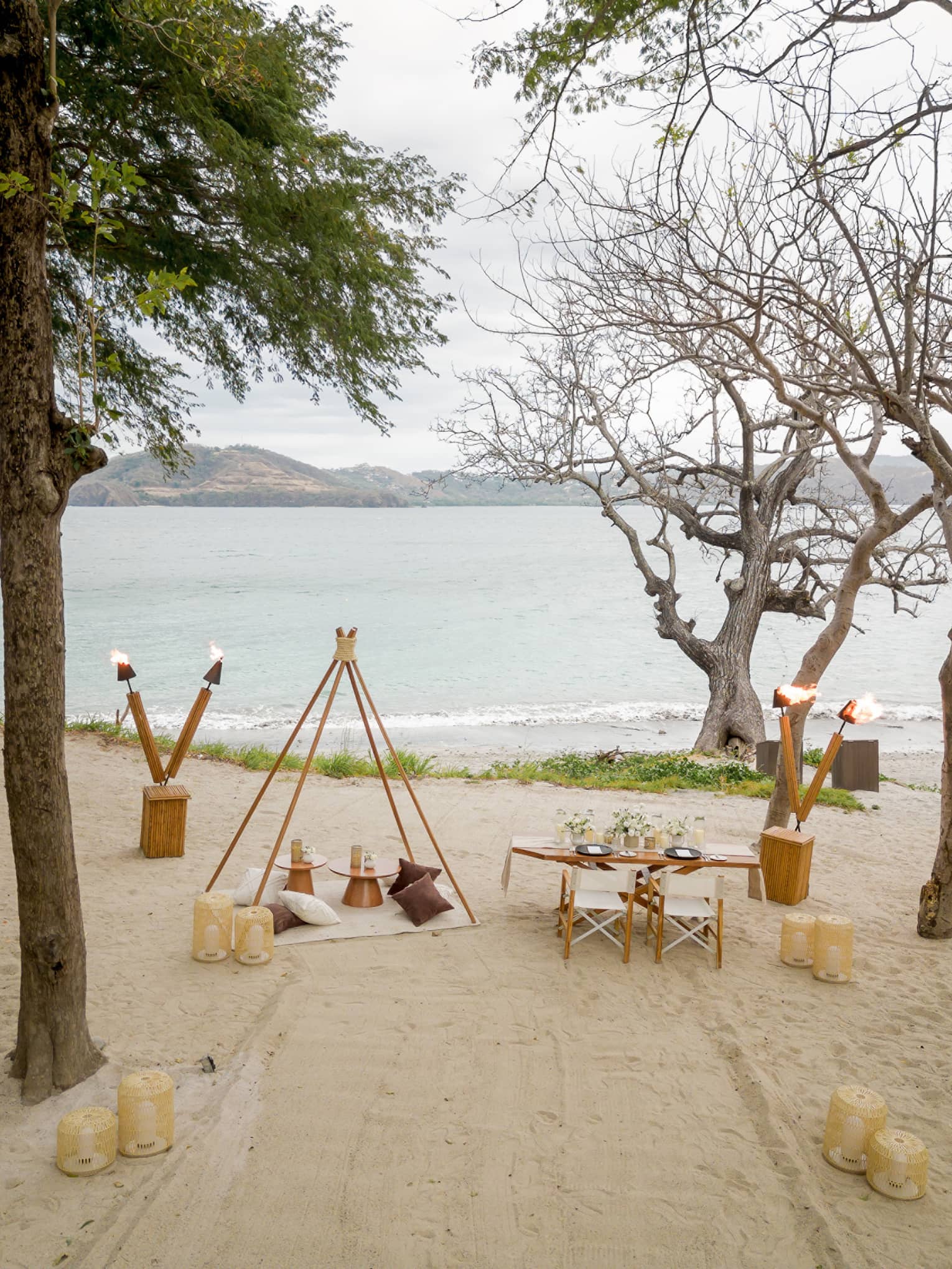 Private beach picnic set up with small table, blanket and pillows, lanterns, and torches
