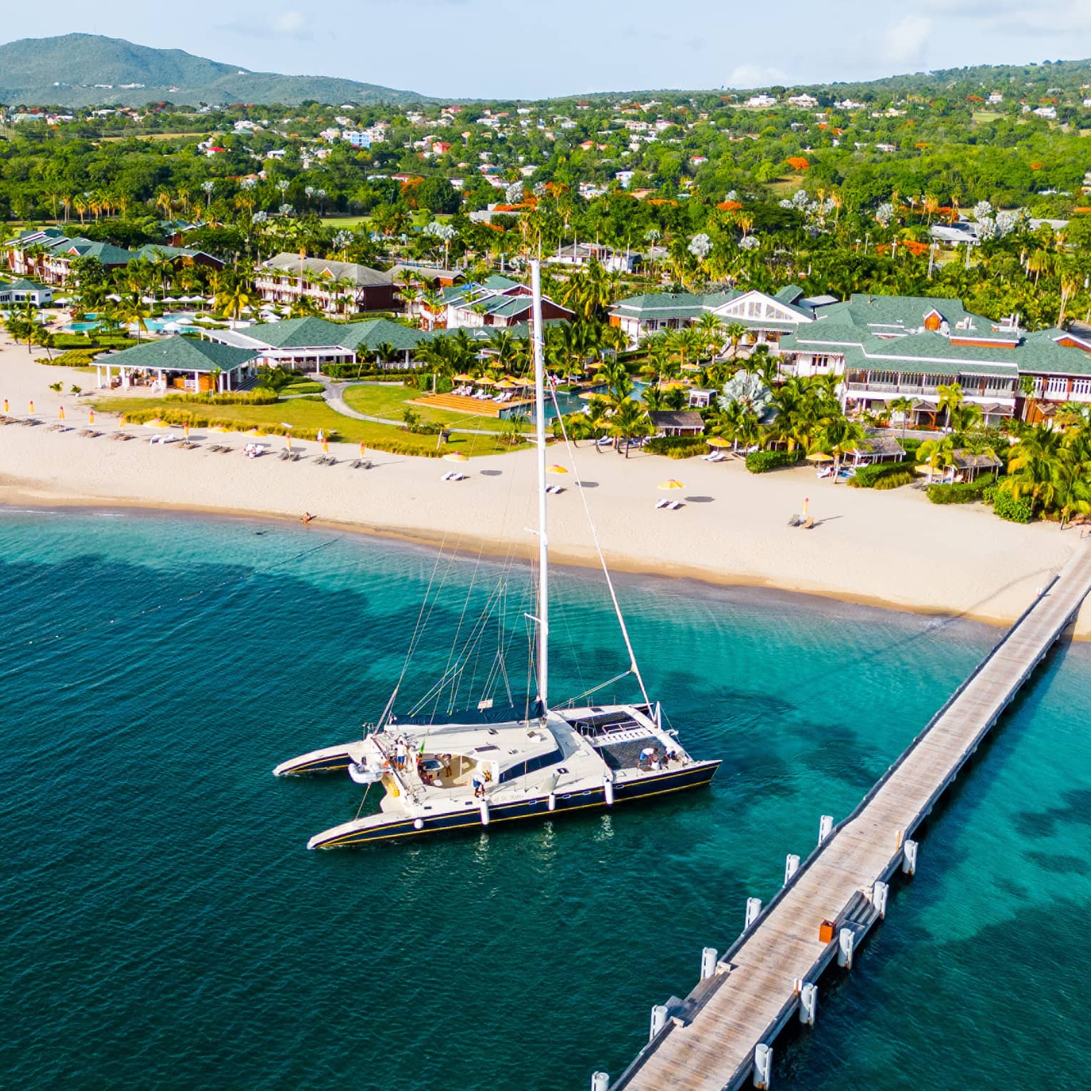 Aerial view of catamaran anchored near a dock reaching out from a beach with a sprawling resort along the coastline set among the trees