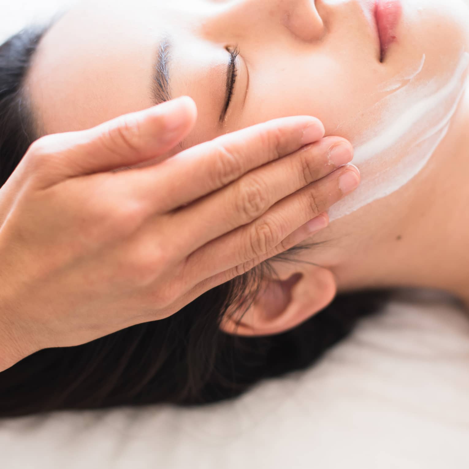 Close-up of spa attendant's hands rubbing white cream on woman's face as she lies on massage table