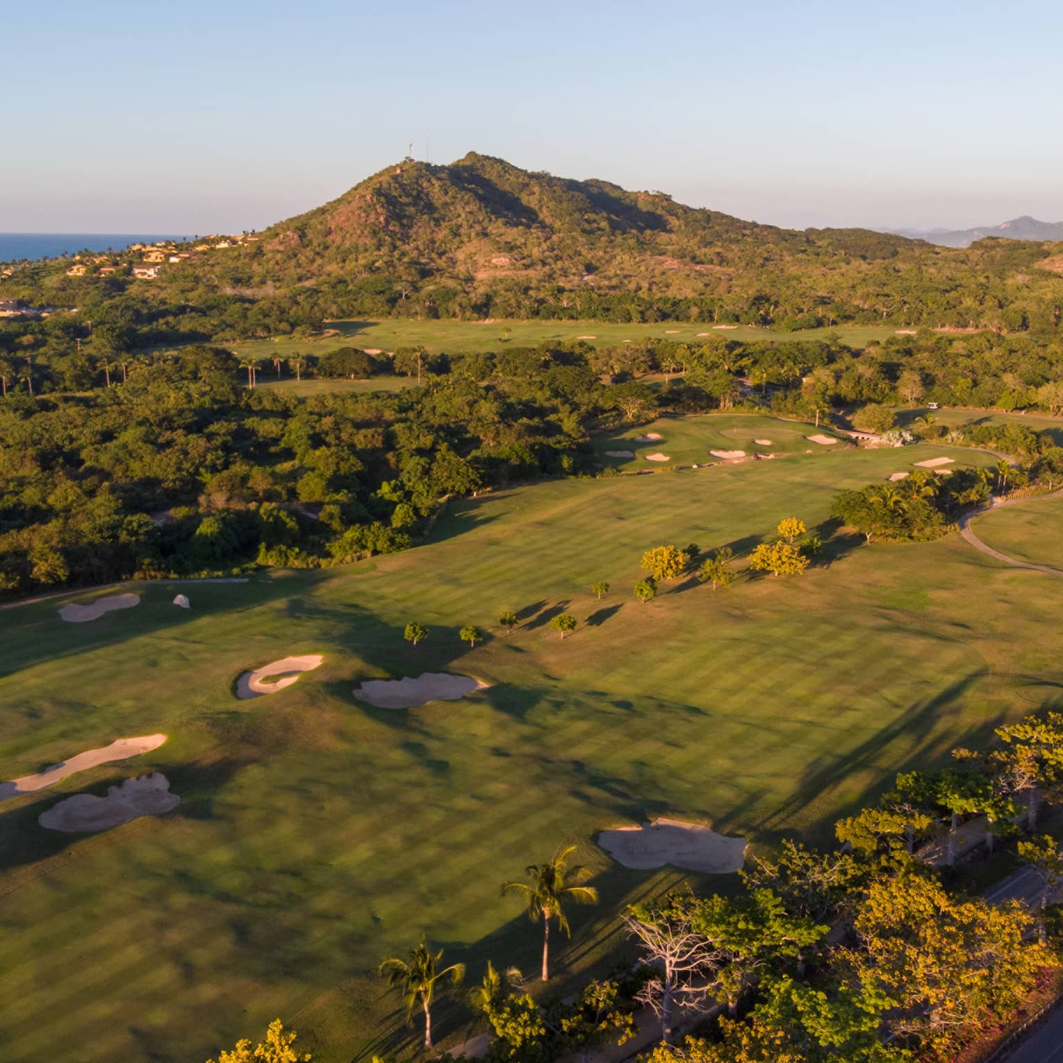 Aerial view of a golf course over the rolling coastal landscape; houses rising along the hillside, ocean in the distance.