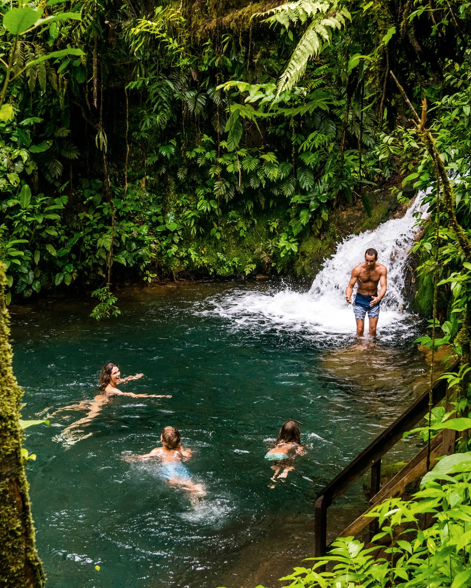 Two adults and two kids frolic at the foot of a waterfall surrounded by lush jungle greenery