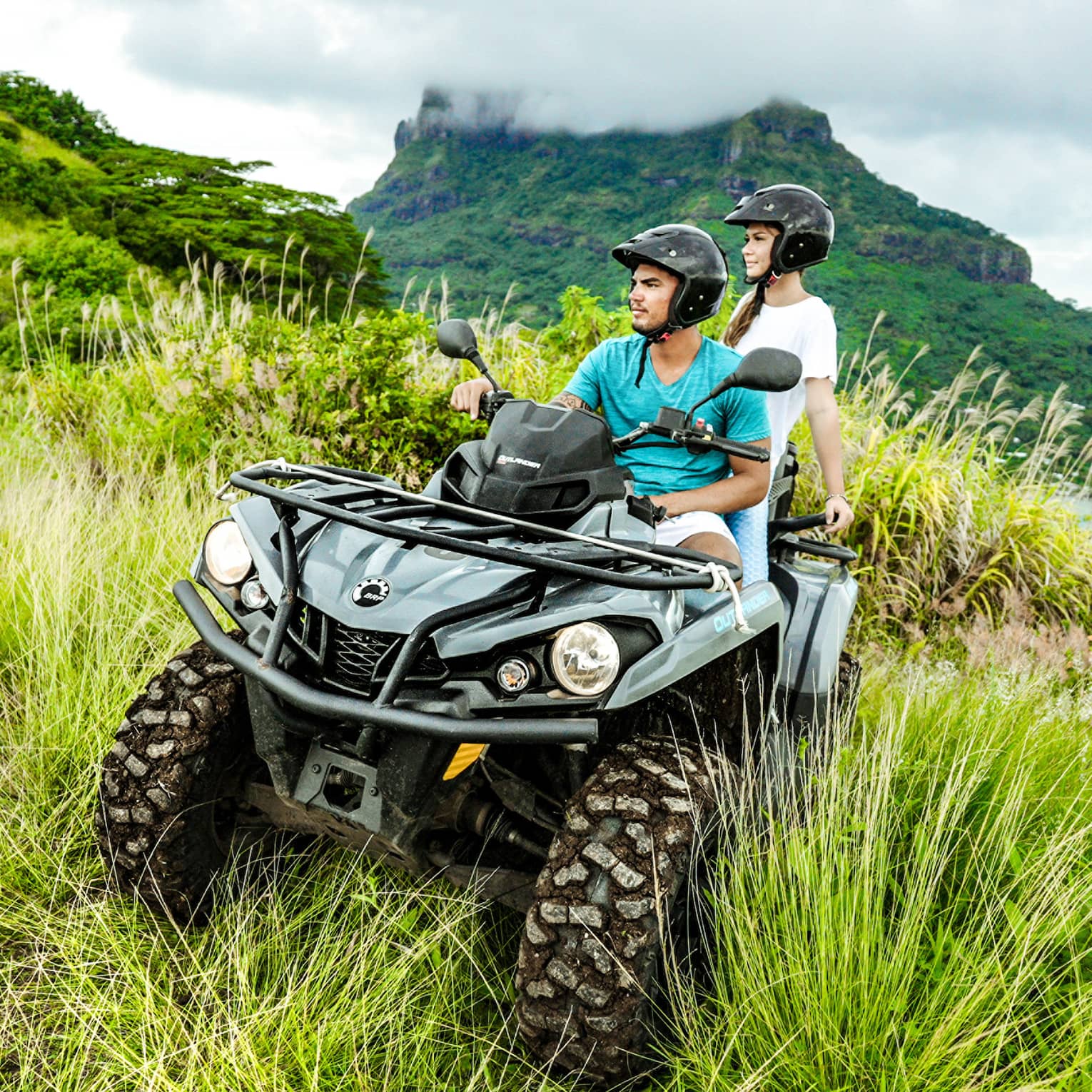 Close-up of a couple riding an ATV through lush vegetation; behind them, a plume of smoke billows above a volcanic mountain.