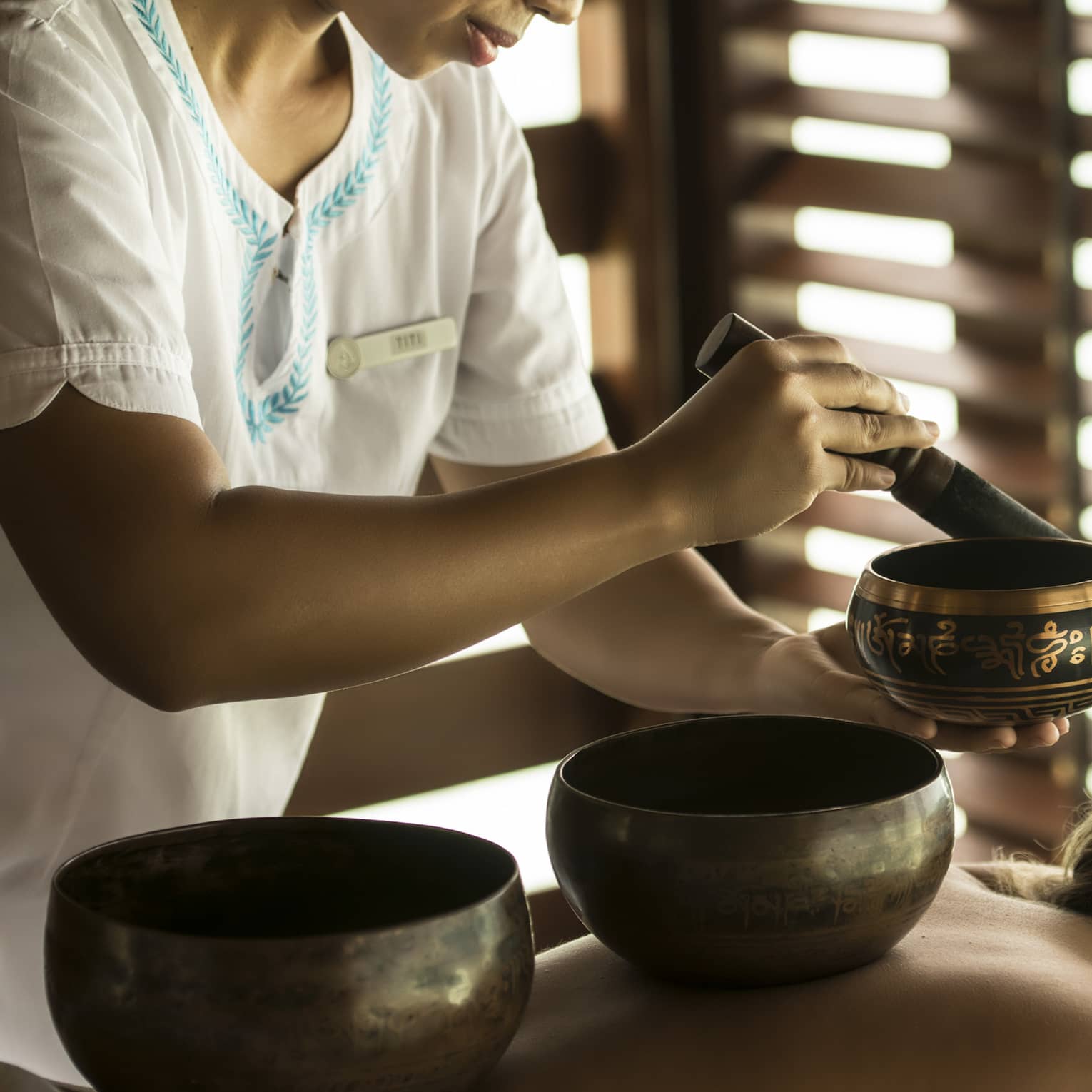 Guest with singing bowls on bare back lies on treatment table which sound specialist plays another singing bowl during a sound healing session