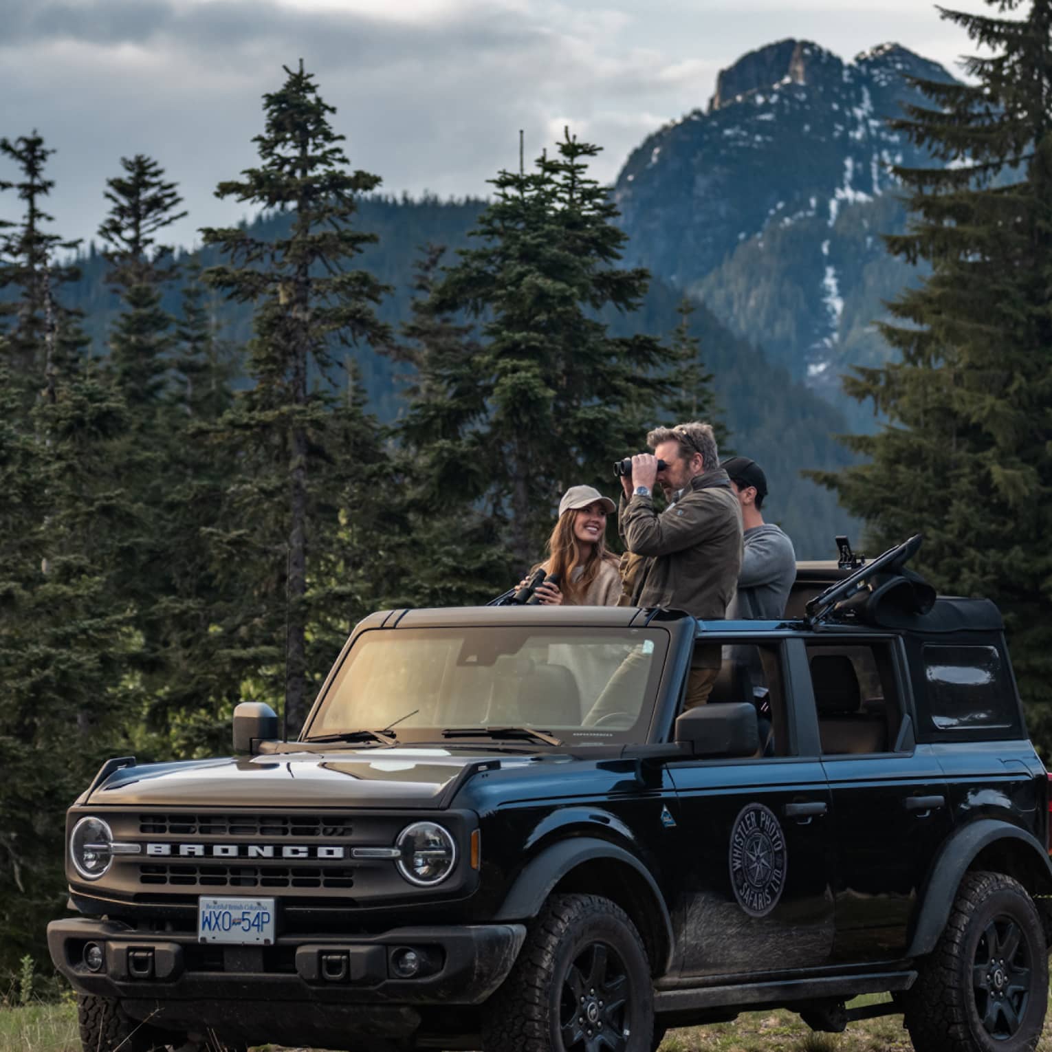 Group of friends with binoculars stand in open-top Ford Bronco surrounded by trees and mountains