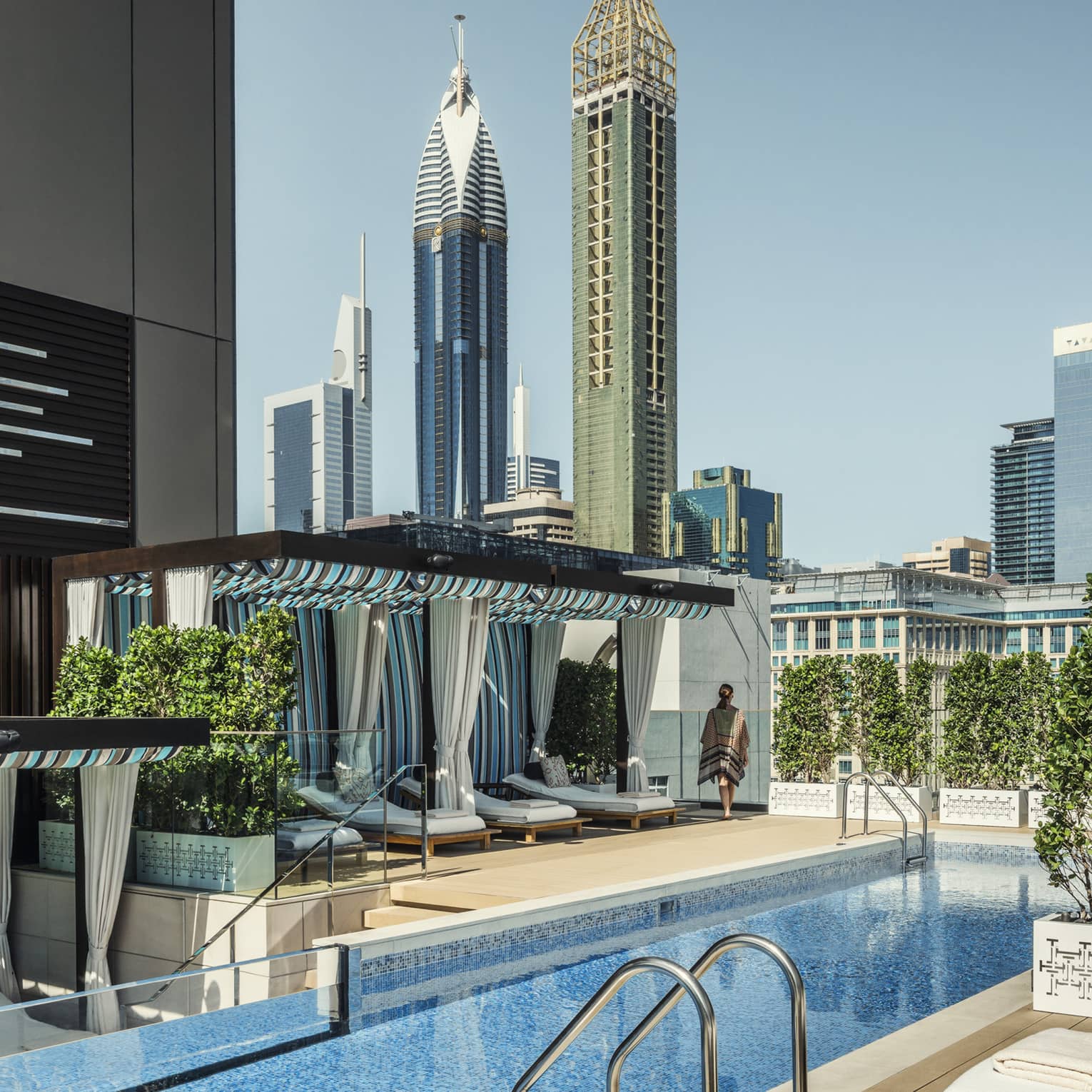 White lounge chairs, ladder into long blue swimming pool by cabanas on rooftop patio on sunny day