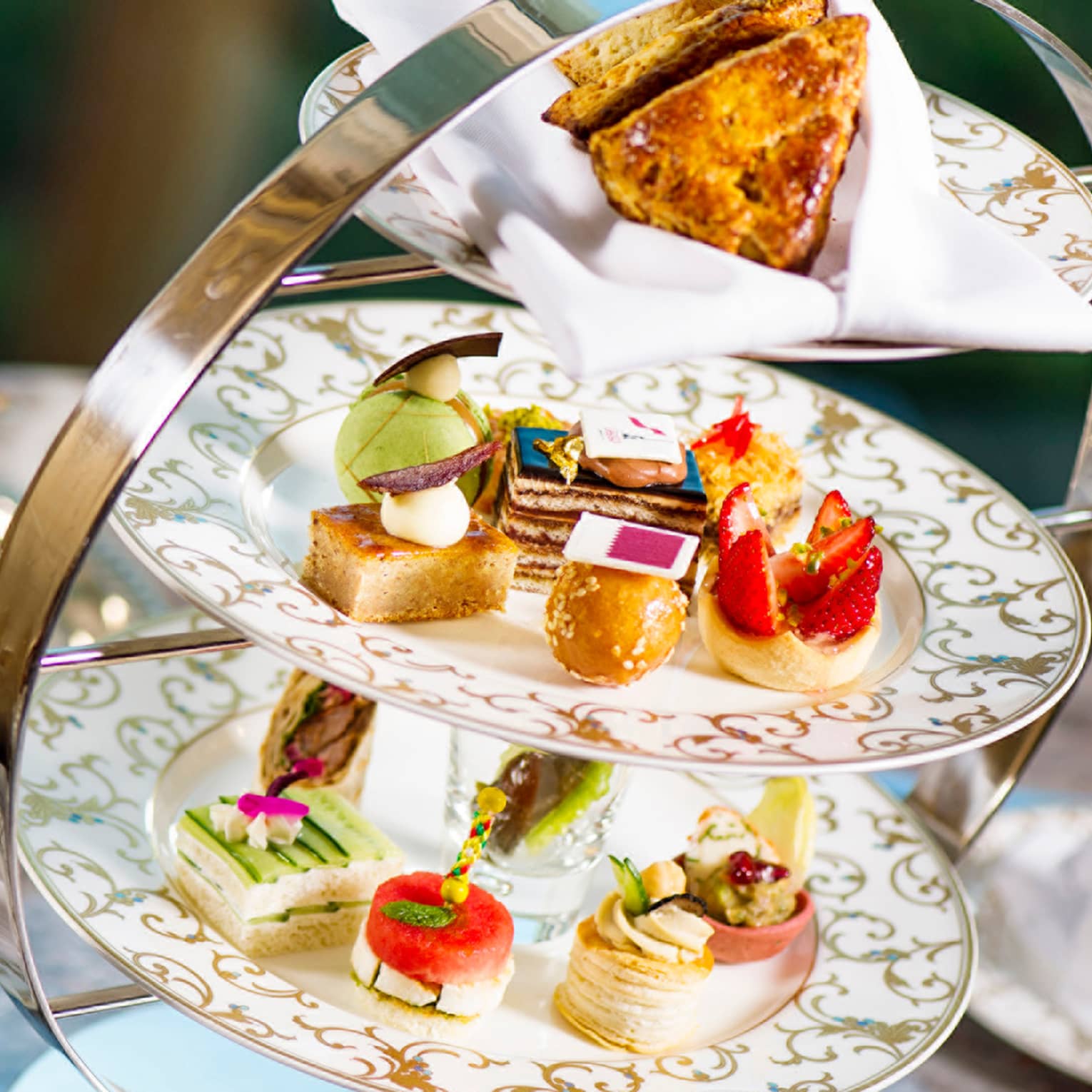 Small, artful pastries displayed on gold-accented china plates in a three-tier stand. Behind, a silver teapot and tray.