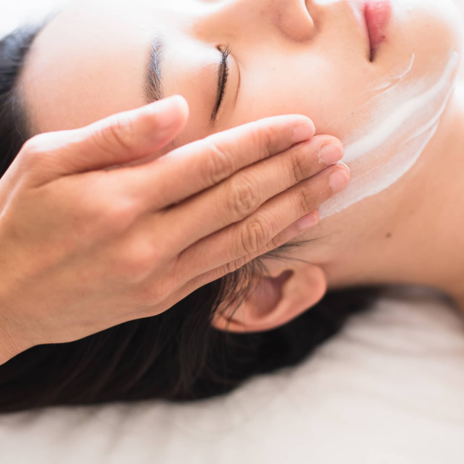 Close-up of spa attendant's hands rubbing white cream on woman's face as she lies on massage table