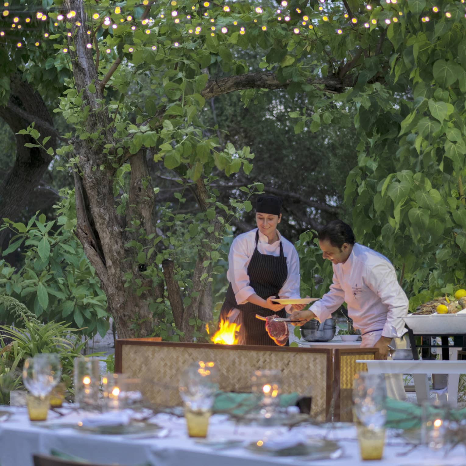 Chefs put large steak on flaming grill by long outdoor dining table, patio lights at night