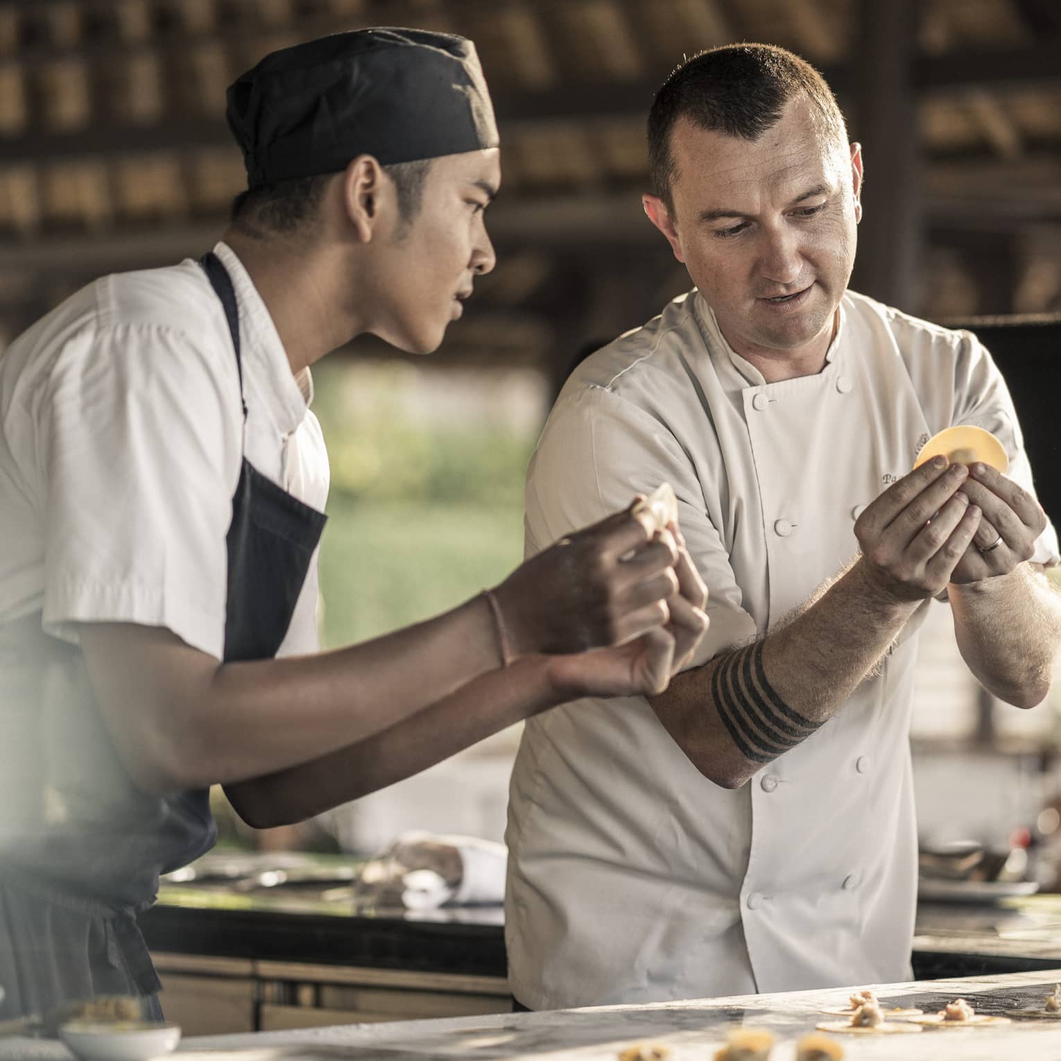A chef teaching a student how to make pasta, folding dough with filling into the shape of a tortellini, in a sunlit kitchen.