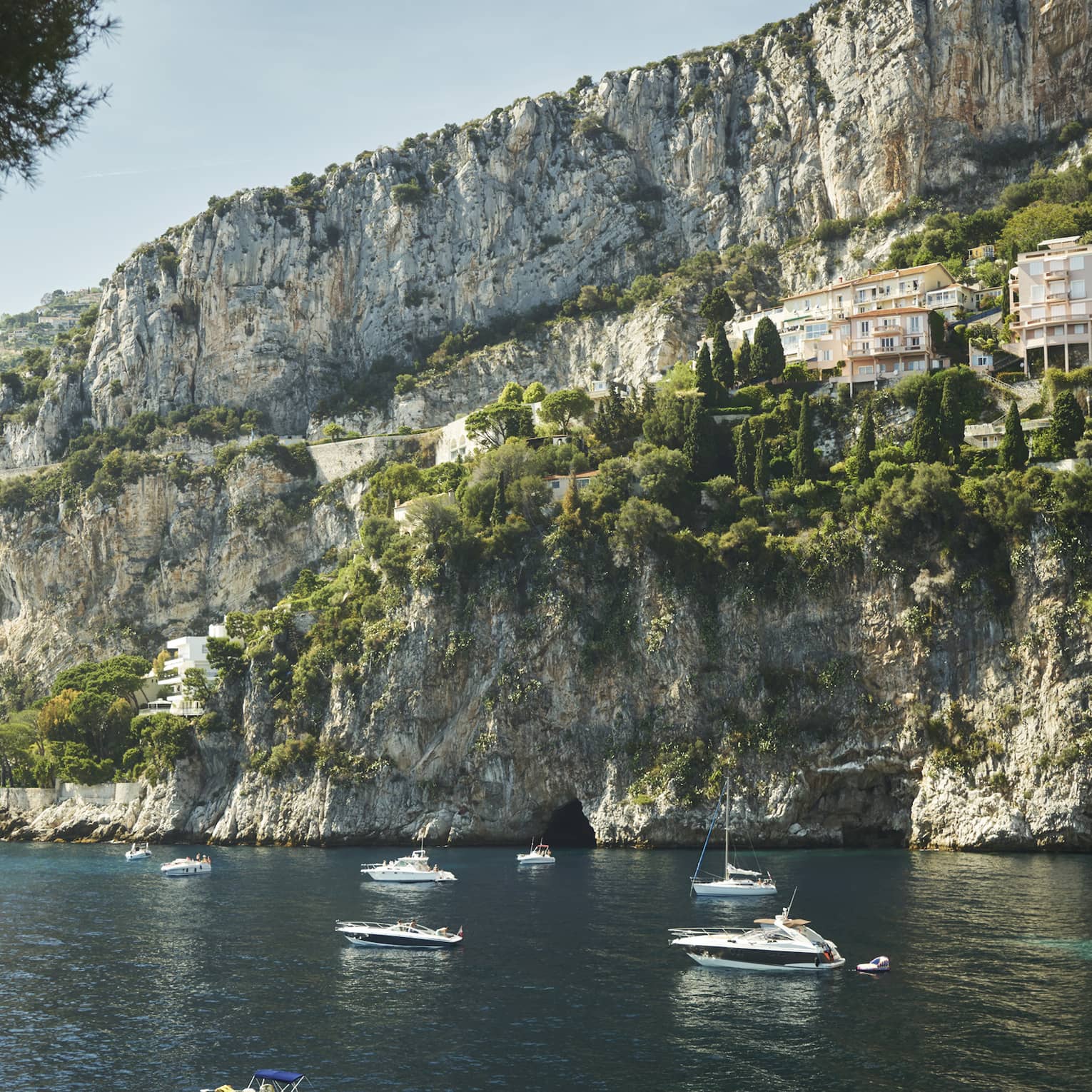Sailboats and yachts float on inky blue water beside a towering cliff face dotted with green trees and pale square buildings.