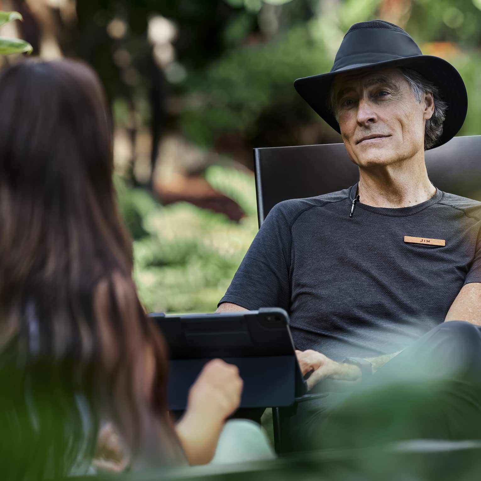 Sitting amid lush vegetation, two people engage in conversation, one with kind eyes and a soft smile typing on a tablet.