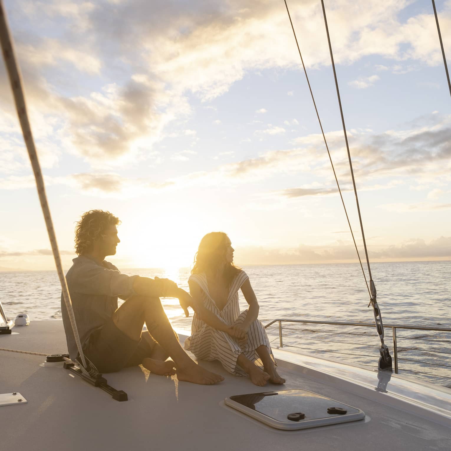 Couple in silhouette sitting barefoot on a boat deck framed by rigging ropes against a bright blue clouded sky at sunset.