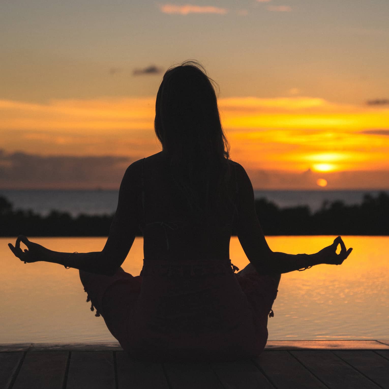 Silhouette of woman in meditation pose in front of calm ocean and orange sunset sky