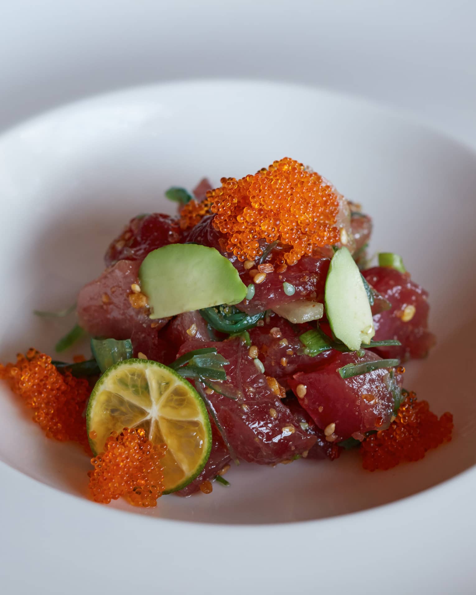 Close-up of tuna sashimi pieces garnished with sesame seeds, avocado, a slice of lime and orange fish roe in a white bowl.