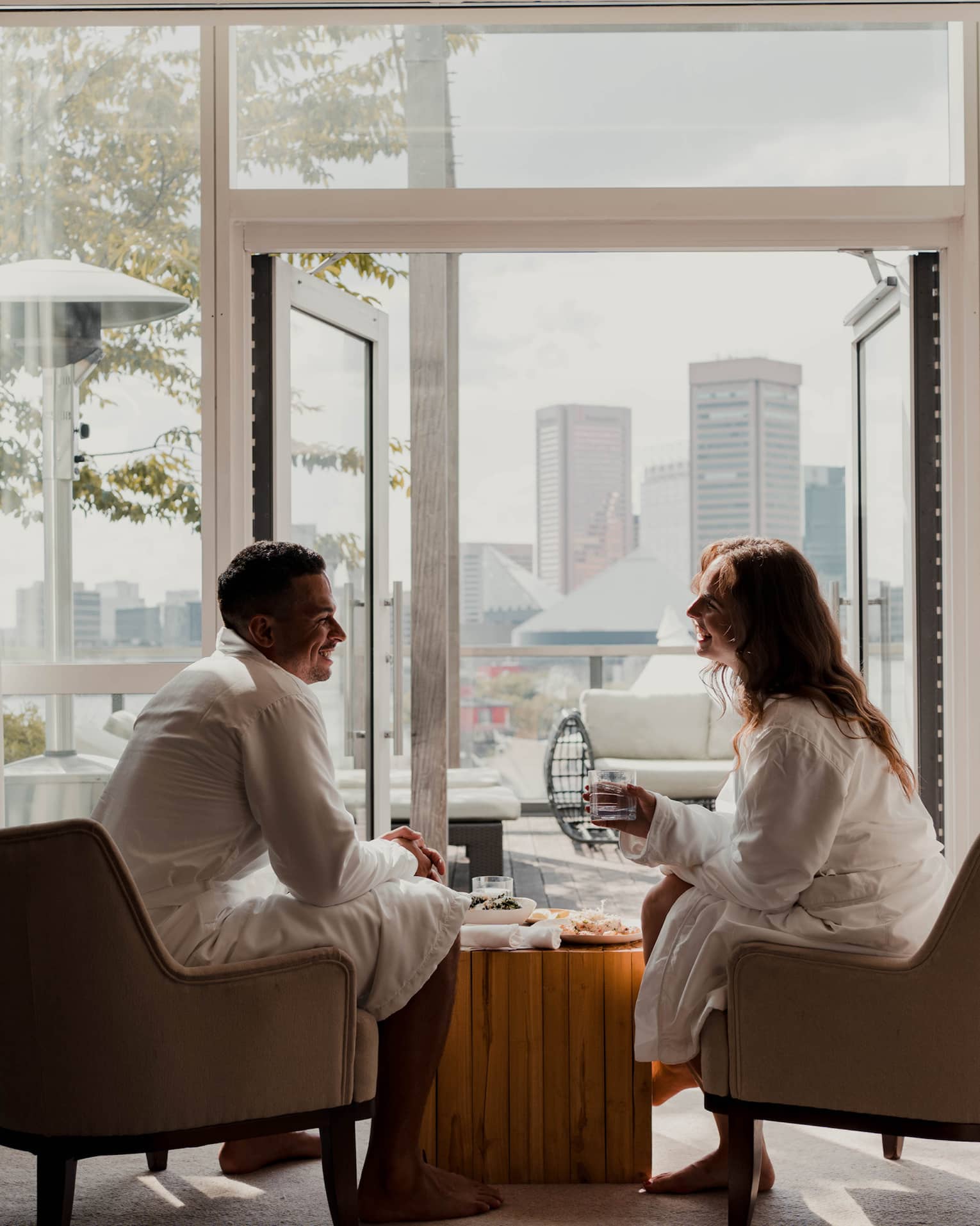 Two people in white robes sit in armchairs by an open balcony door with a cityscape view in the background, sharing a relaxed, casual conversation.