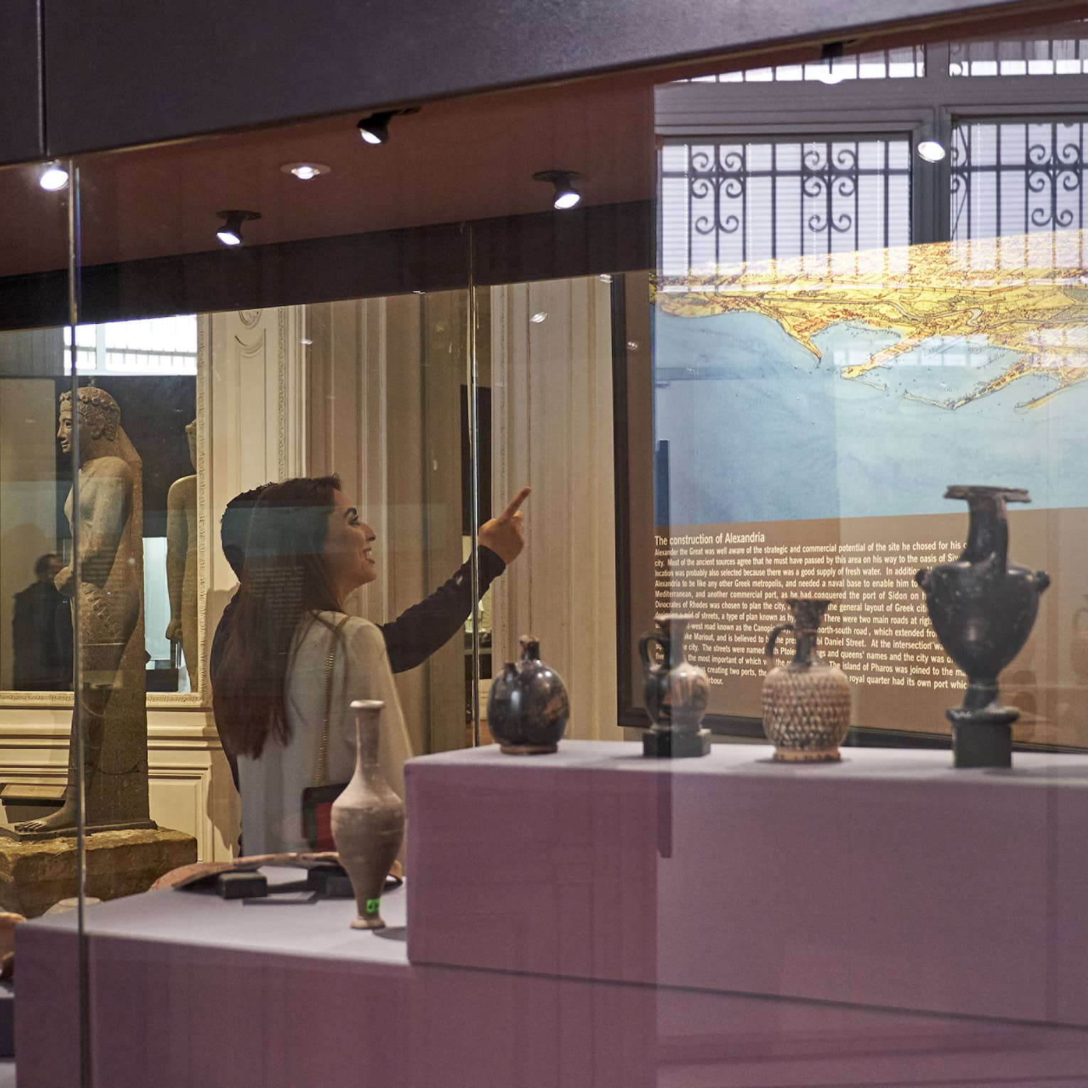 A visitor in a museum points up at a historical map lit up on a wall, behind them a display case with various vases and jugs.