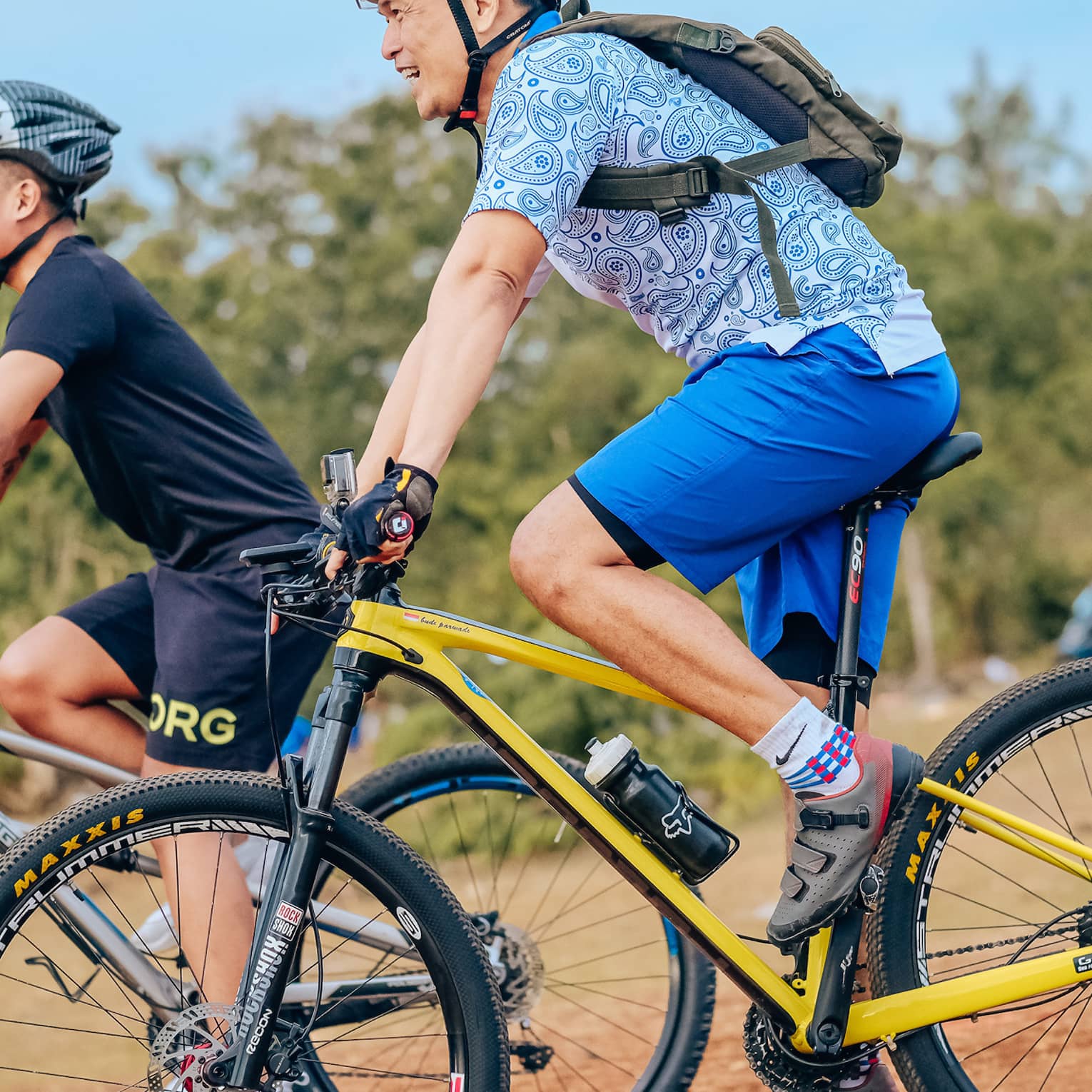 Side view of two smiling mountain bikers in helmets as they coast down a gentle slope on a dirt path alongside trees.