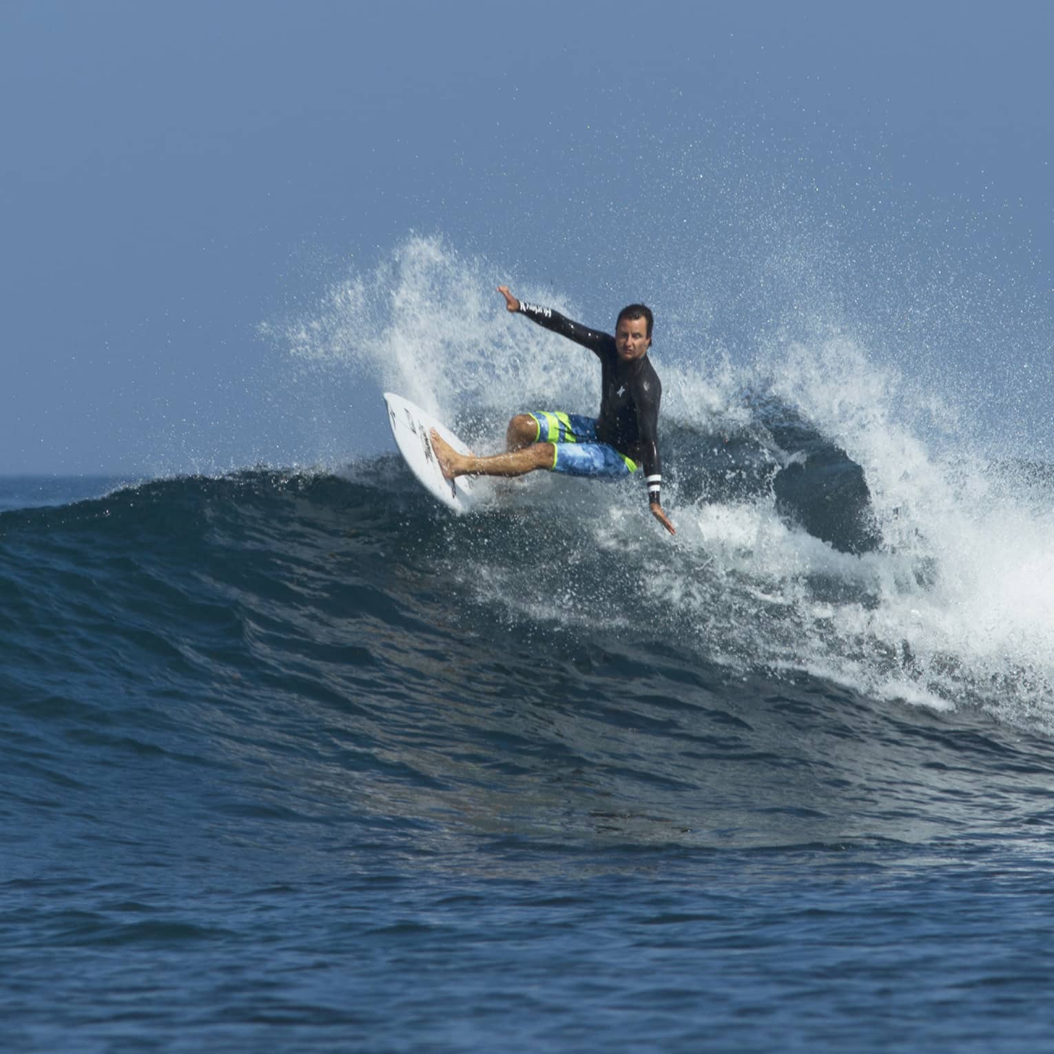 A surfer in a long sleeve rash guard and board shorts rides a wave, arms outstretched, in the wake of frothy white spray.