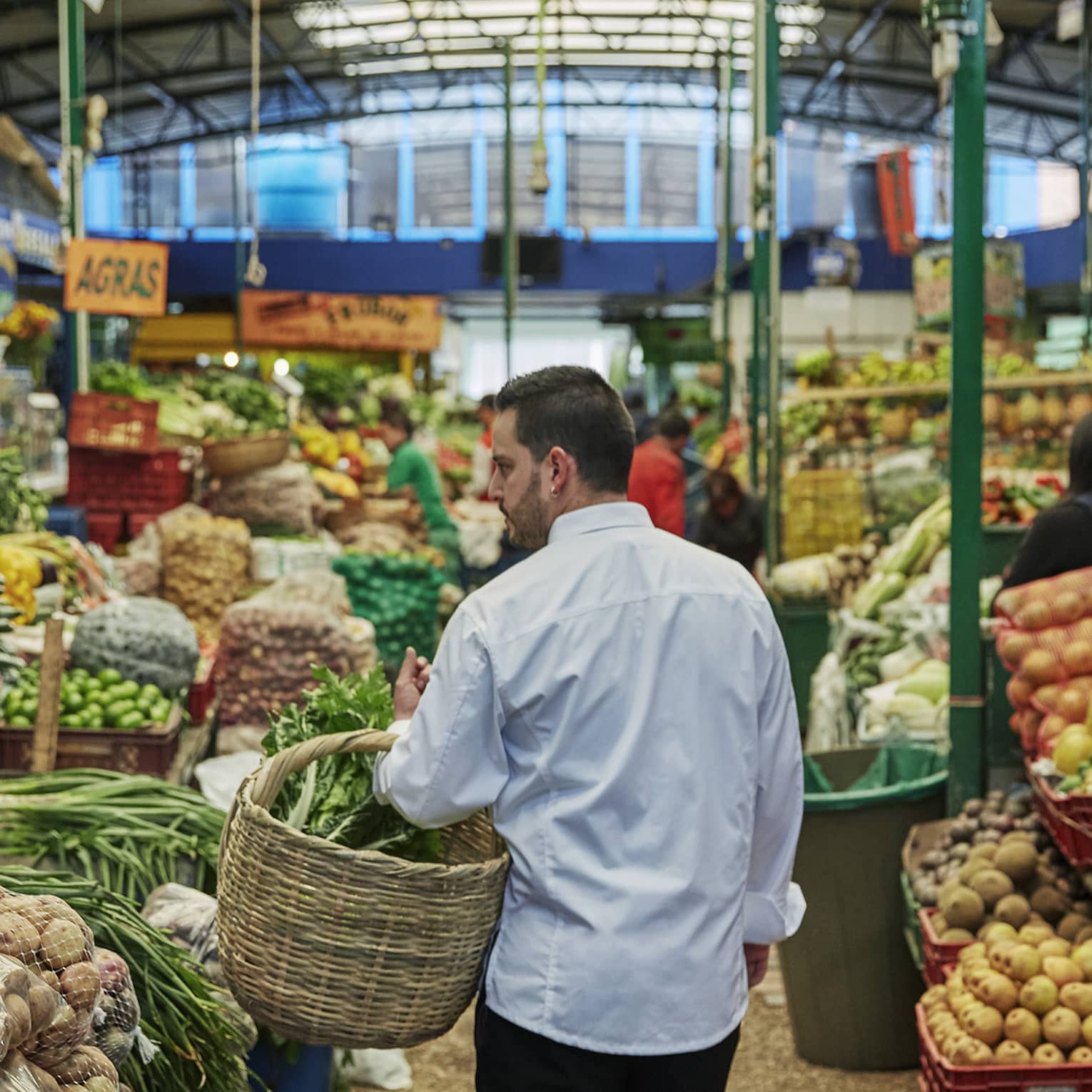 Back of man with beard carrying wicker basket through fruit market aisle, produce stacked on shelves