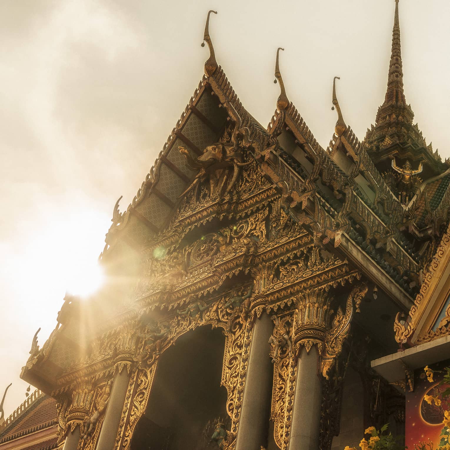 Intricately designed temple with ornate carvings and tiered roofs, illuminated by sunlight, with a tree and yellow flowers in the foreground
