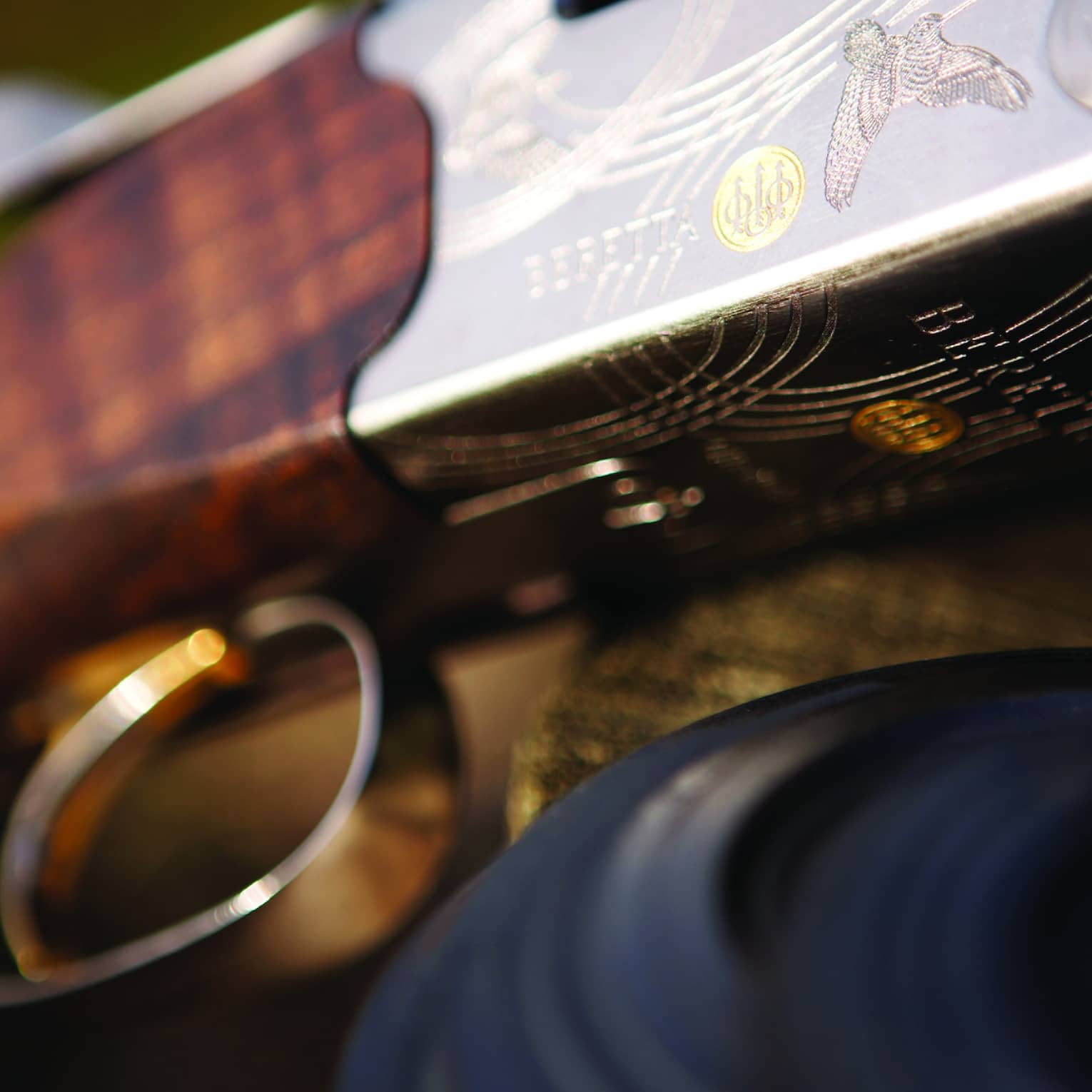 Close-up of trigger and wooden barrel of a rifle with silver engraving of a grouse, the Beretta name, swirls and a gold logo.