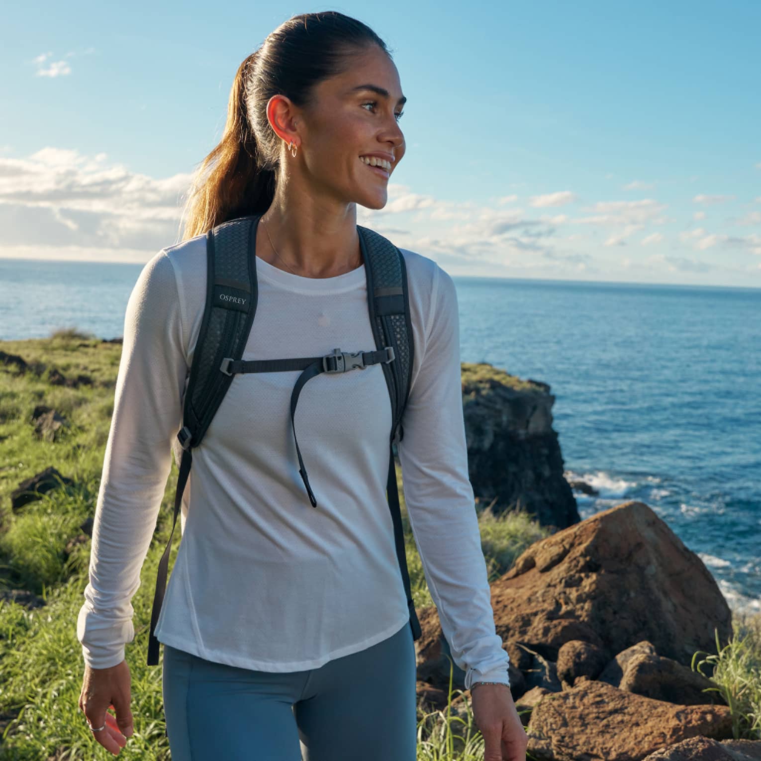Close-up of a smiling, backpacked guest standing atop a grassy coastal cliff, clear sky and ocean with gentle wake beyond.