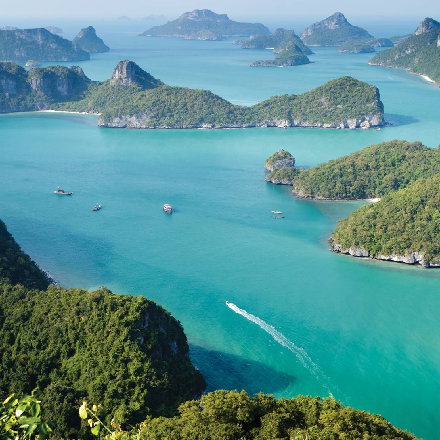 Aerial view of a dozen lush, forested, hilly islands of different sizes surrounded by calm turquoise water dotted with boats.