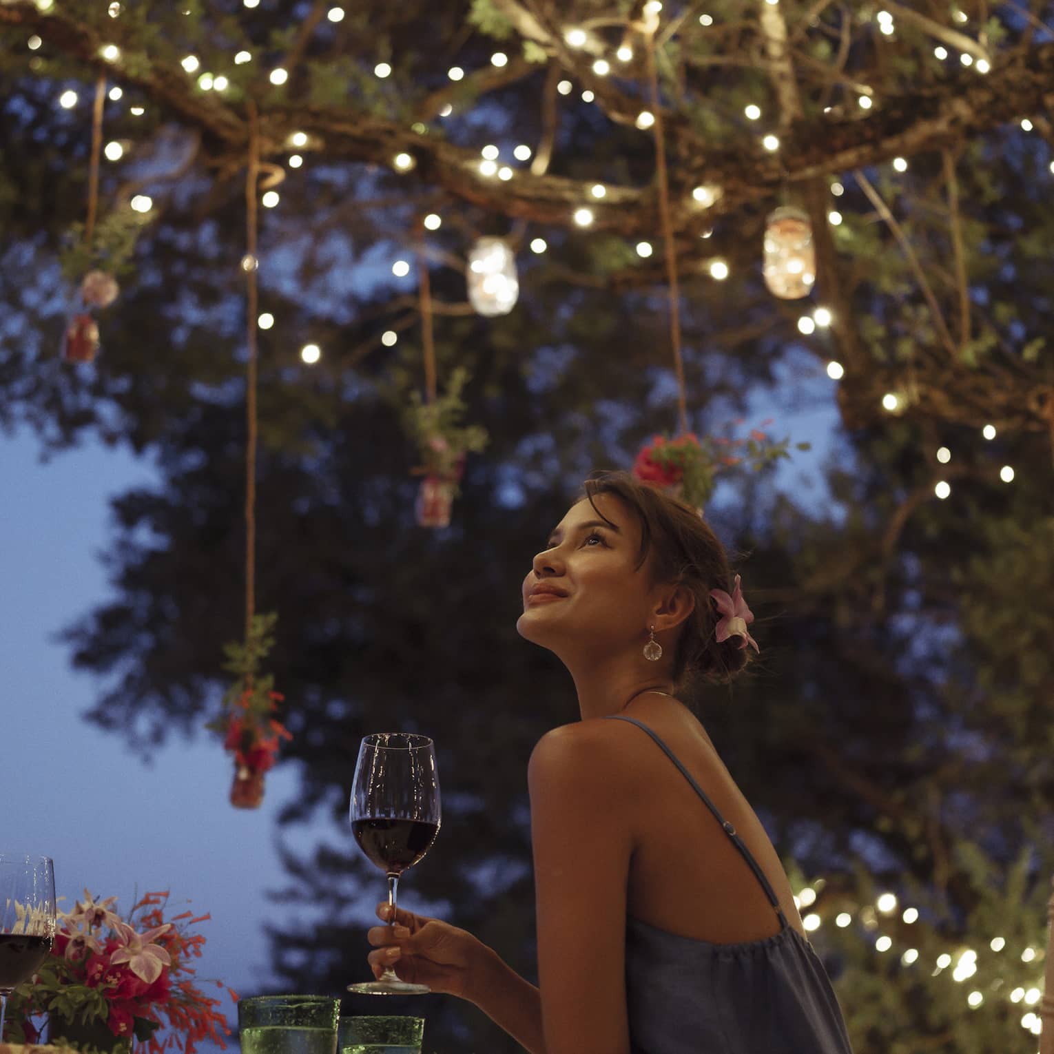 A smiling couple having a romantic dinner under trees covered in string lights and jars of flowers hanging from the branches.