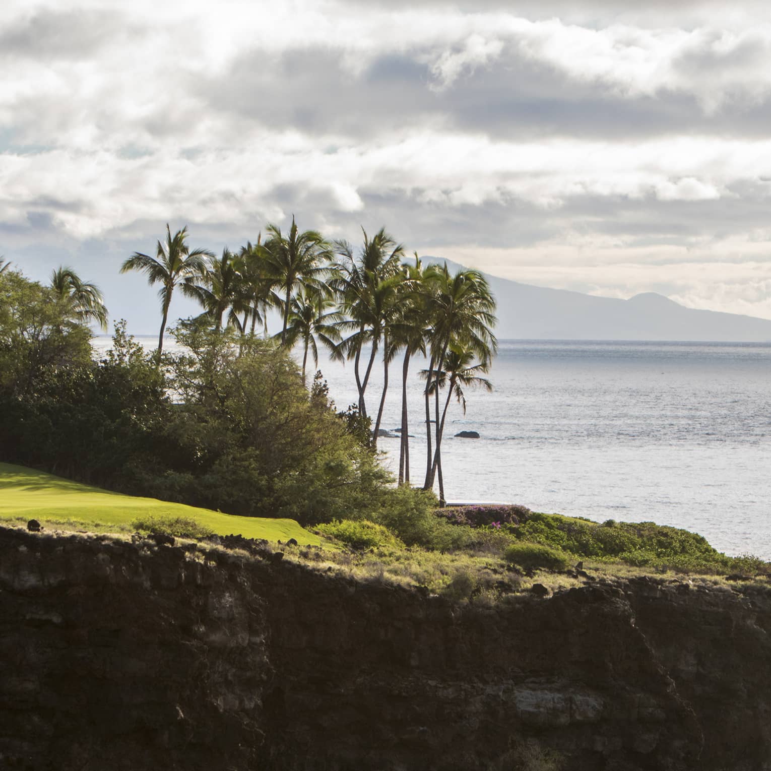 Long view of two people on clifftop golf green aside lush trees and towering palms; fluffy clouds, ocean and mountain beyond.
