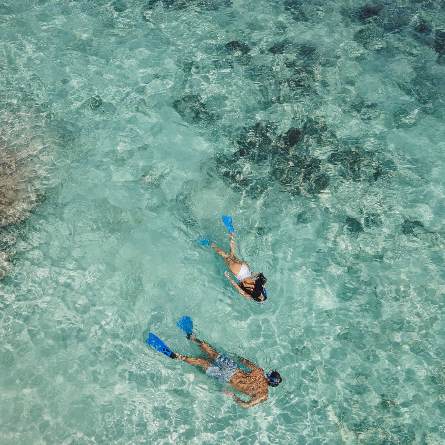 Aerial view of two snorkellers wearing blue flippers in an expanse of shallow, turquoise waters rippling over the reef.