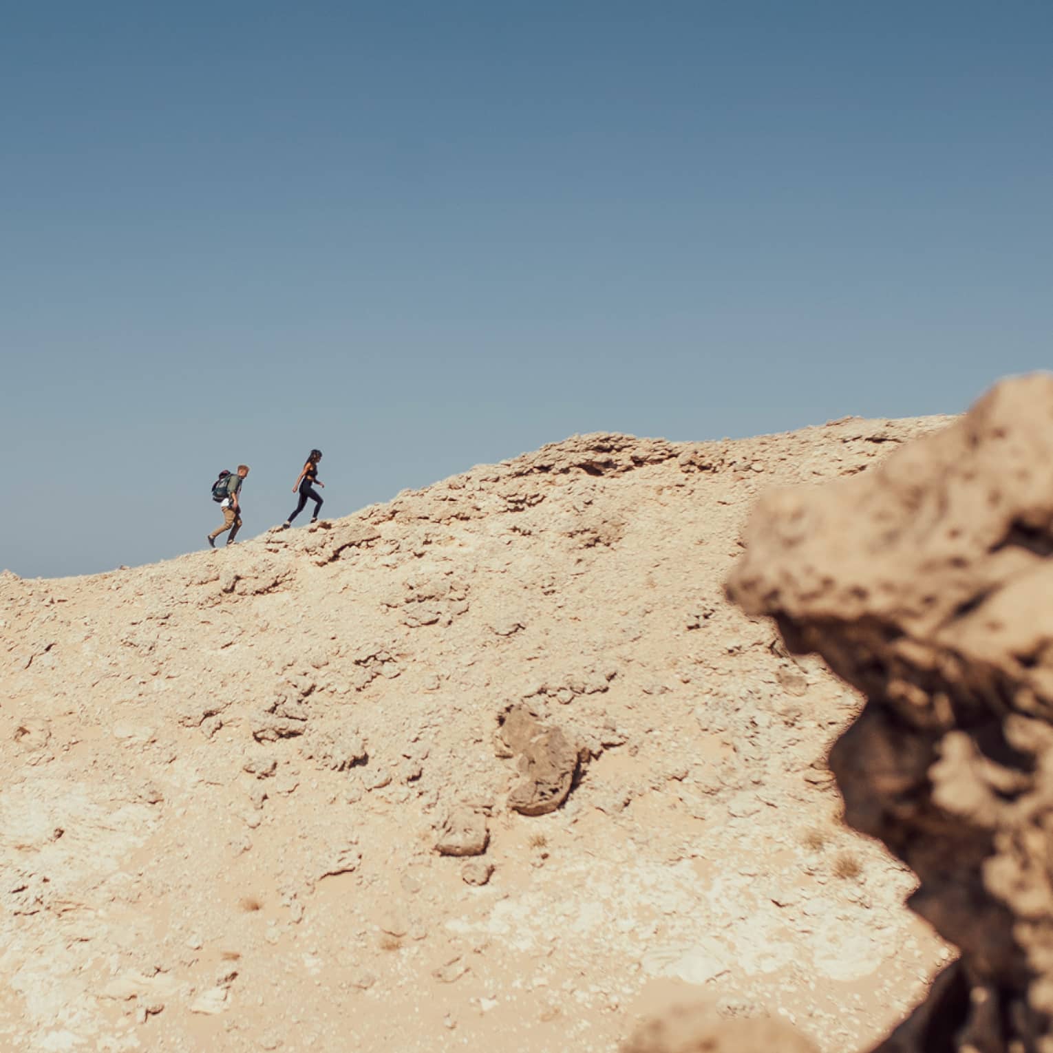Against a background of sunny, clear blue sky and surrounded by rocky terrain, two people hike up a sandy hillside.