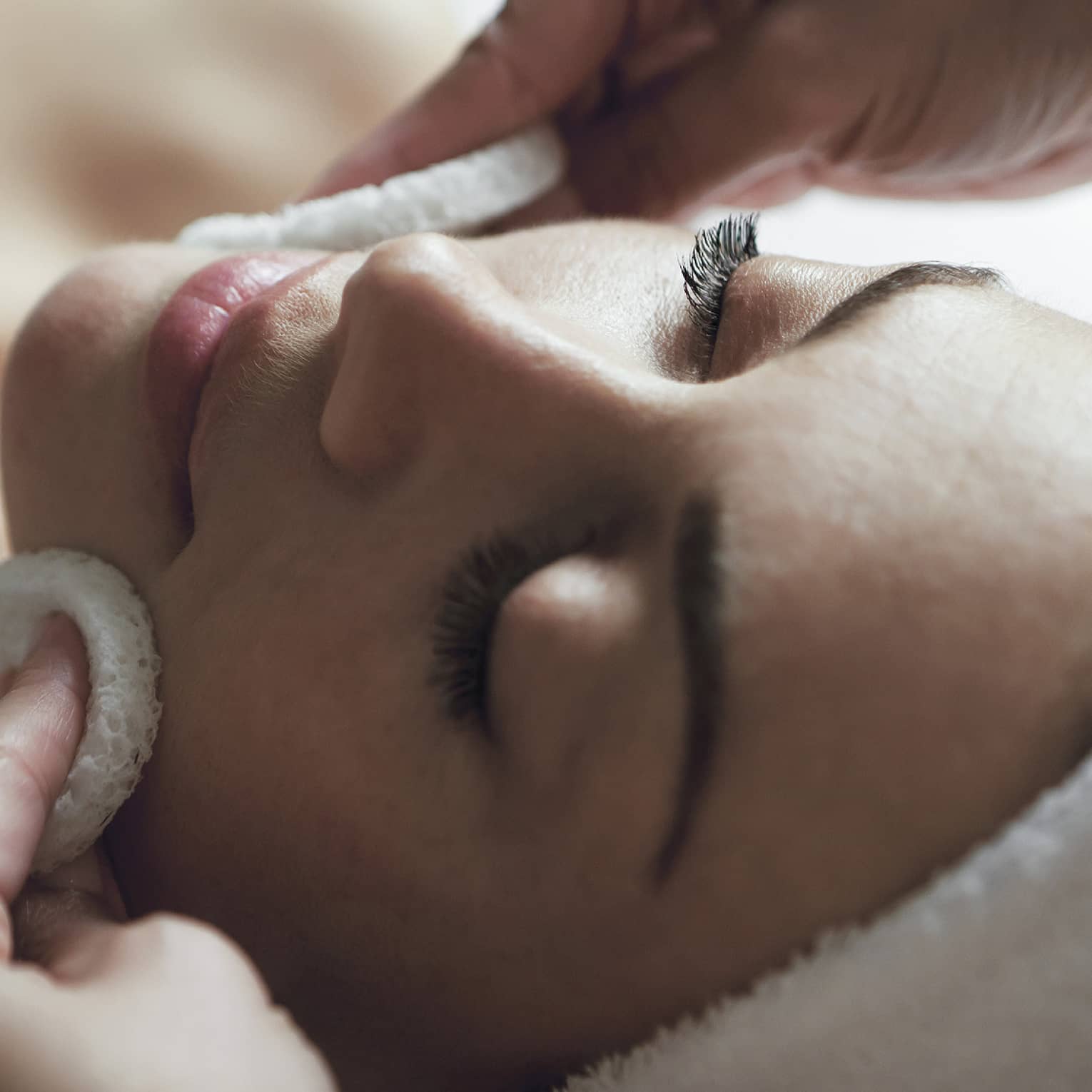 A detail of hands rubbing a sponge on a woman's face as she lies on a massage table with her eyes closed and hair wrapped up in a white towel
