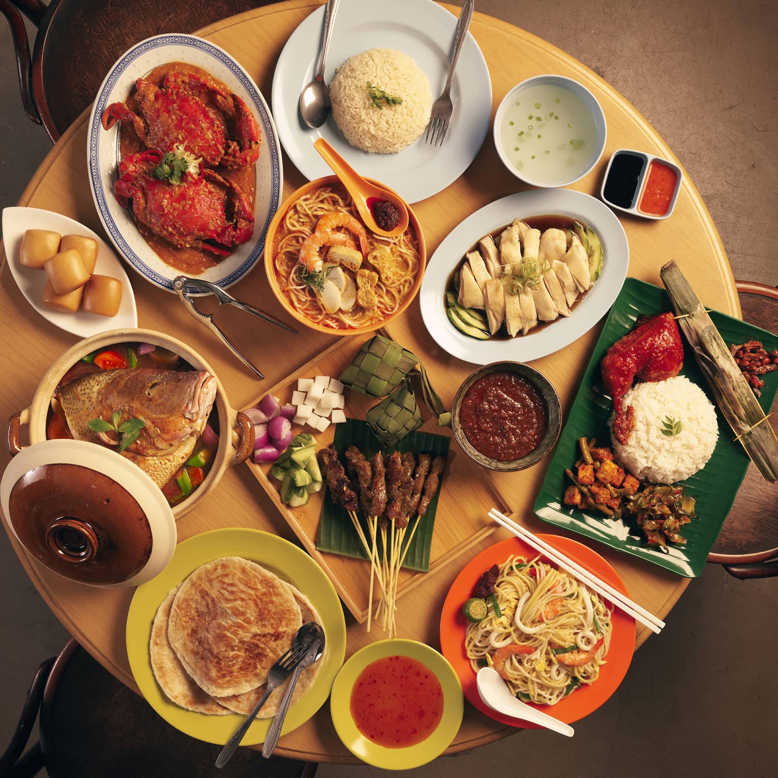 Overhead view of a round table covered in colourful dishes of crab, prawns, chicken, noodles, rice, skewers and flatbread